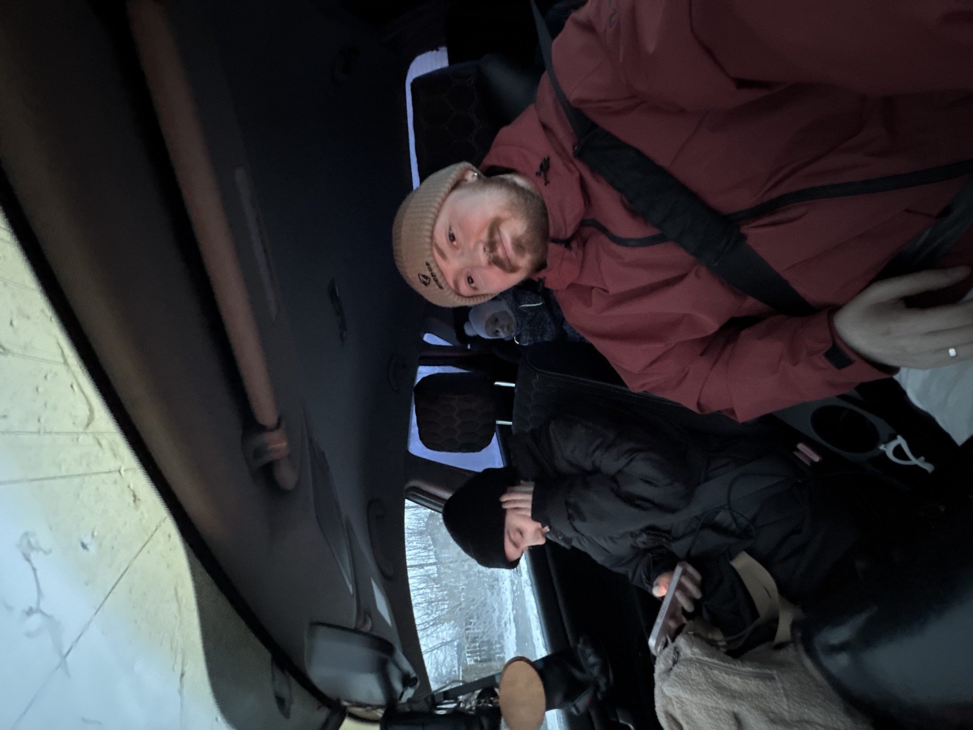 Two individuals are inside a car. One person is wearing a red jacket with a seatbelt, and a beanie. The other person is wearing a black jacket and a beanie. There is a phone in view. The photograph was taken in Chaikovsky, Russia. The landscape outside suggests it might be winter.