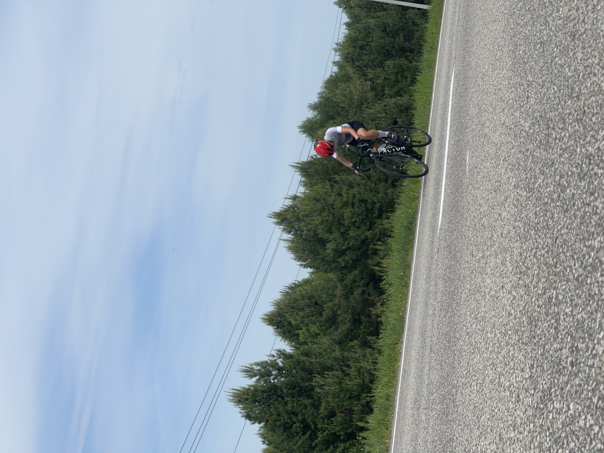 The image depicts a cyclist riding on a paved road. The cyclist is wearing a red helmet, white shirt, and dark shorts. The bicycle is black and has the word "FACTOR" printed on the side. The road is gray and has a white line marking the edge. The cyclist is positioned on the right side of the road. To the left of the road is a grassy area followed by a line of trees. The sky is light blue with faint clouds. The location is potentially Maksimovo, Russia.