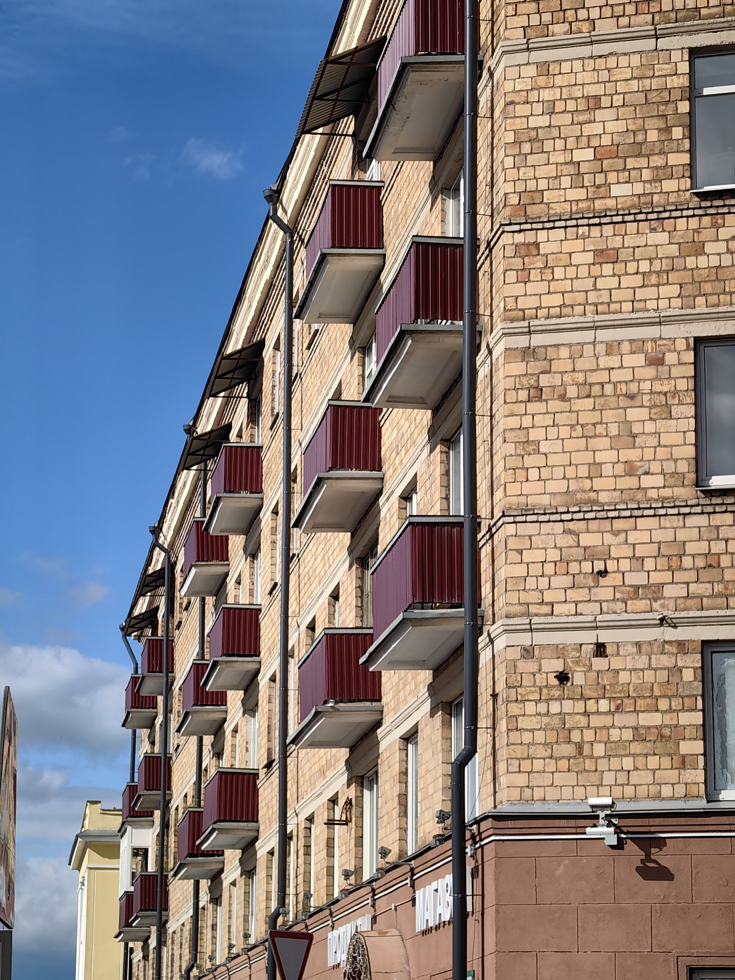 This is a close-up, low-angle shot of a building facade, likely an apartment building, in Minsk, Belarus. The building is constructed primarily of light-colored brick with darker reddish-brown trim on the lower section. Several floors are visible, each featuring uniformly designed balconies with dark red metal railings and white floor panels. Black drainpipes run vertically along the side of the building. The upper edge of the building shows a dark metal roofline with several small awnings extending over the balconies.
The sky above is a clear, vibrant blue with a few wispy white clouds, suggesting it is daytime with fair weather. The sunlight casts subtle shadows, indicating the sun is positioned to the side, likely mid-morning or afternoon.
In the foreground, on the lower right side, a red triangular traffic sign is partially visible. To the left of this sign, the building's lower facade is covered in larger, reddish-brown tiles. A white sign with dark text, possibly a shop name, is mounted on this lower section, with the visible portion reading "МАГАЗ". A surveillance camera is mounted on the wall above this sign.
To the far left of the image, the yellow facade of an adjacent building is visible, hinting at an urban streetscape. The overall impression is one of a typical urban environment with residential and commercial elements, under a bright, clear sky. There are no people visible in the image.