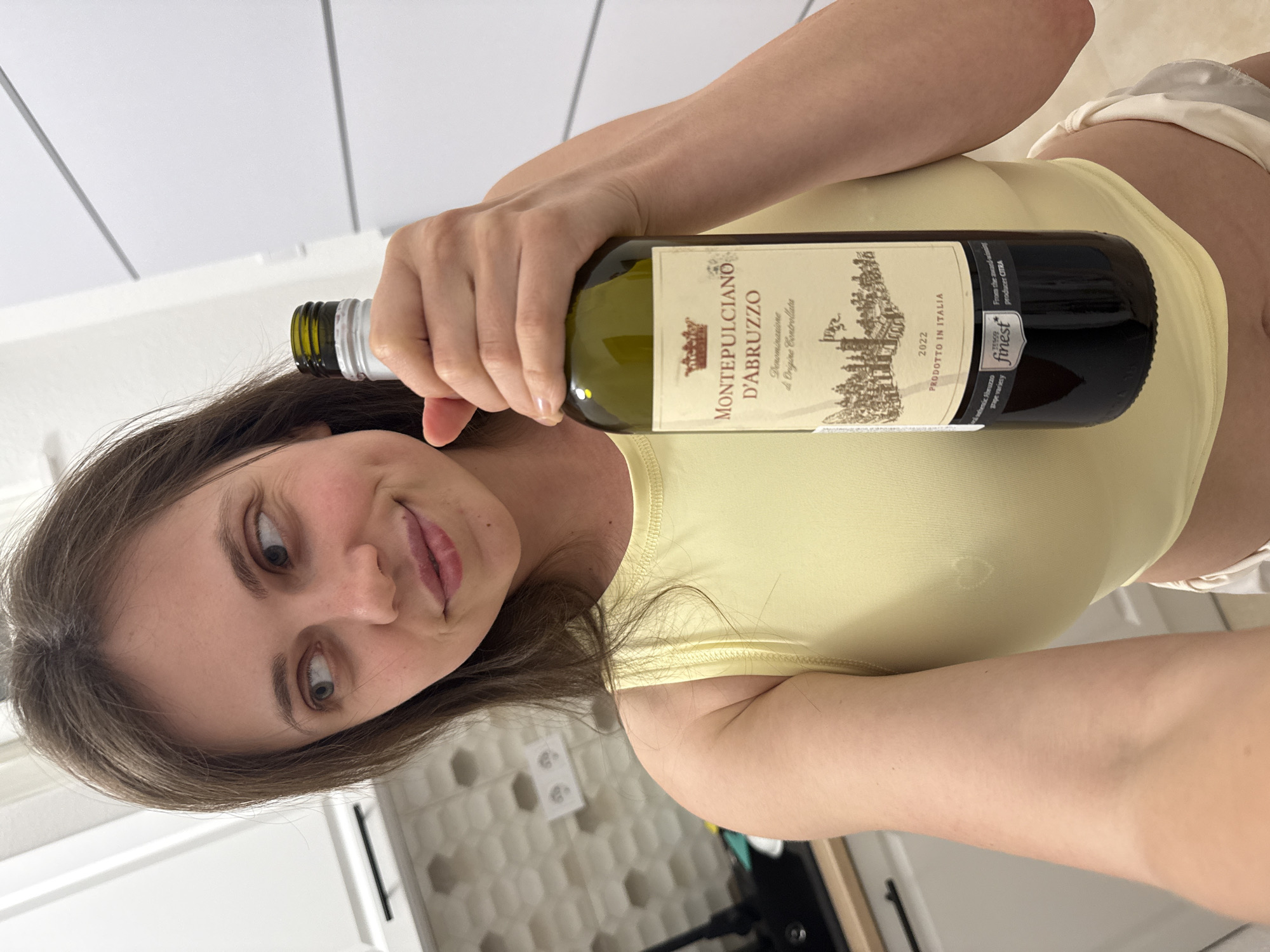 A woman is holding a bottle of "Montepulciano d'Abruzzo" wine in what appears to be a kitchen setting. The wine label indicates it's a 2022 vintage, "Prodotto in Italia" (Product of Italy), and sourced from the award-winning producer CITRA, Tesco finest, featuring authentic Abruzzo grape variety. The surroundings include white wall panels, suggesting an interior space, possibly a kitchen in a residential building. There is no information suggesting the location in the city of Kőröshegy, Hungary.