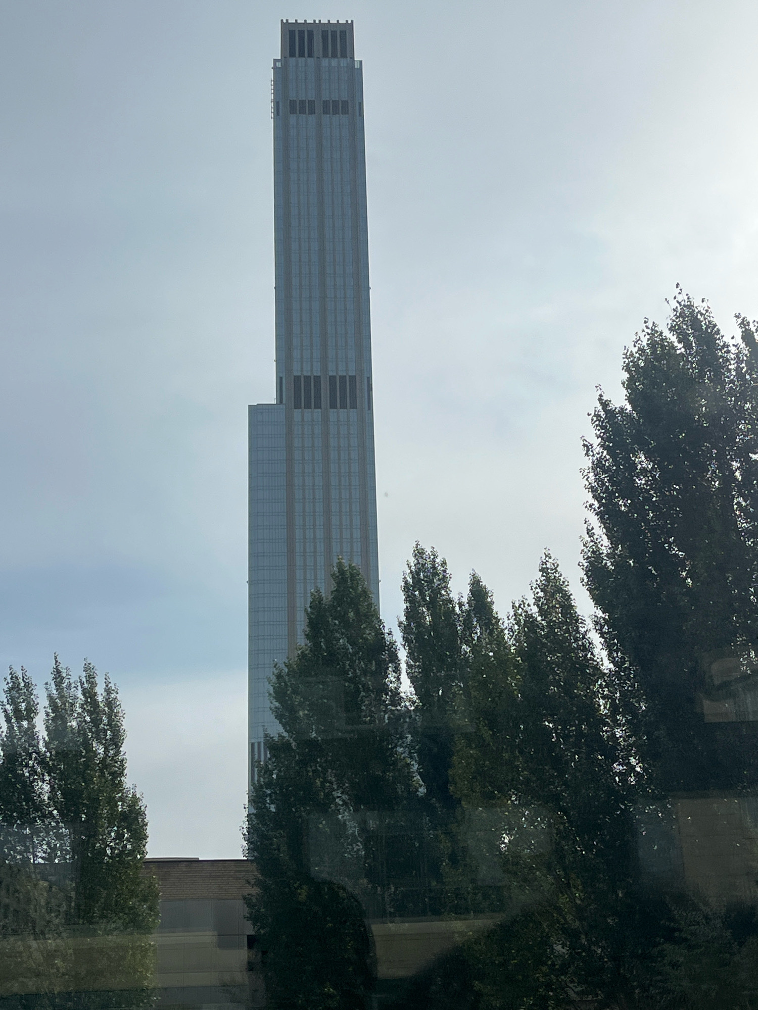 The image shows a very tall, slender skyscraper with a light-blue, glass facade, reaching into a hazy, light-gray sky. The top of the building has a distinct, stepped crown with dark, rectangular windows. Below the crown, there are two tiers of dark, rectangular windows spaced evenly along the facade. The lower portion of the skyscraper is visible, and it appears to be angled or set back slightly. In the foreground and to the sides, a dense cluster of tall, dark green trees with dense foliage partially obscures the lower parts of the scene. A low-rise building with a brick facade and darker windows is visible behind the trees and to the left of the skyscraper. The overall impression is that of an urban landscape on a slightly overcast day. This is the Astana National Bank.