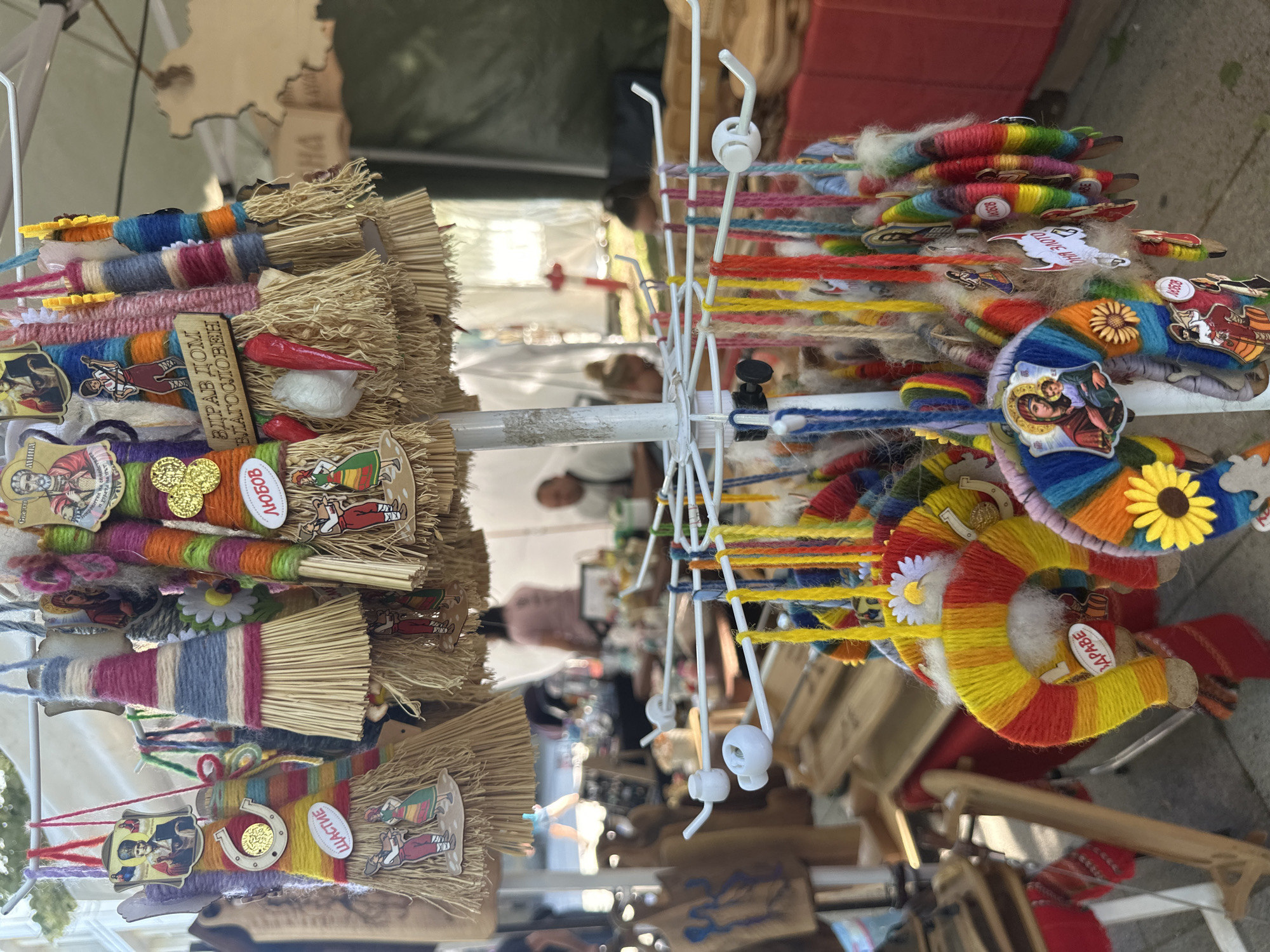 The image depicts a display of handcrafted items, likely for sale, at an outdoor market in Montana, Bulgaria. The objects are predominantly small, colorful brooms or decorative items adorned with thread, fabric, buttons, small religious icons, and miniature figurines. The market setting is suggested by the presence of other stalls and blurred figures in the background. The image is well-lit, indicating daytime. The brooms are mounted on a white, multi-tiered metal display rack with hooks. The colors and materials used in the crafts suggest a folk art or traditional aesthetic.