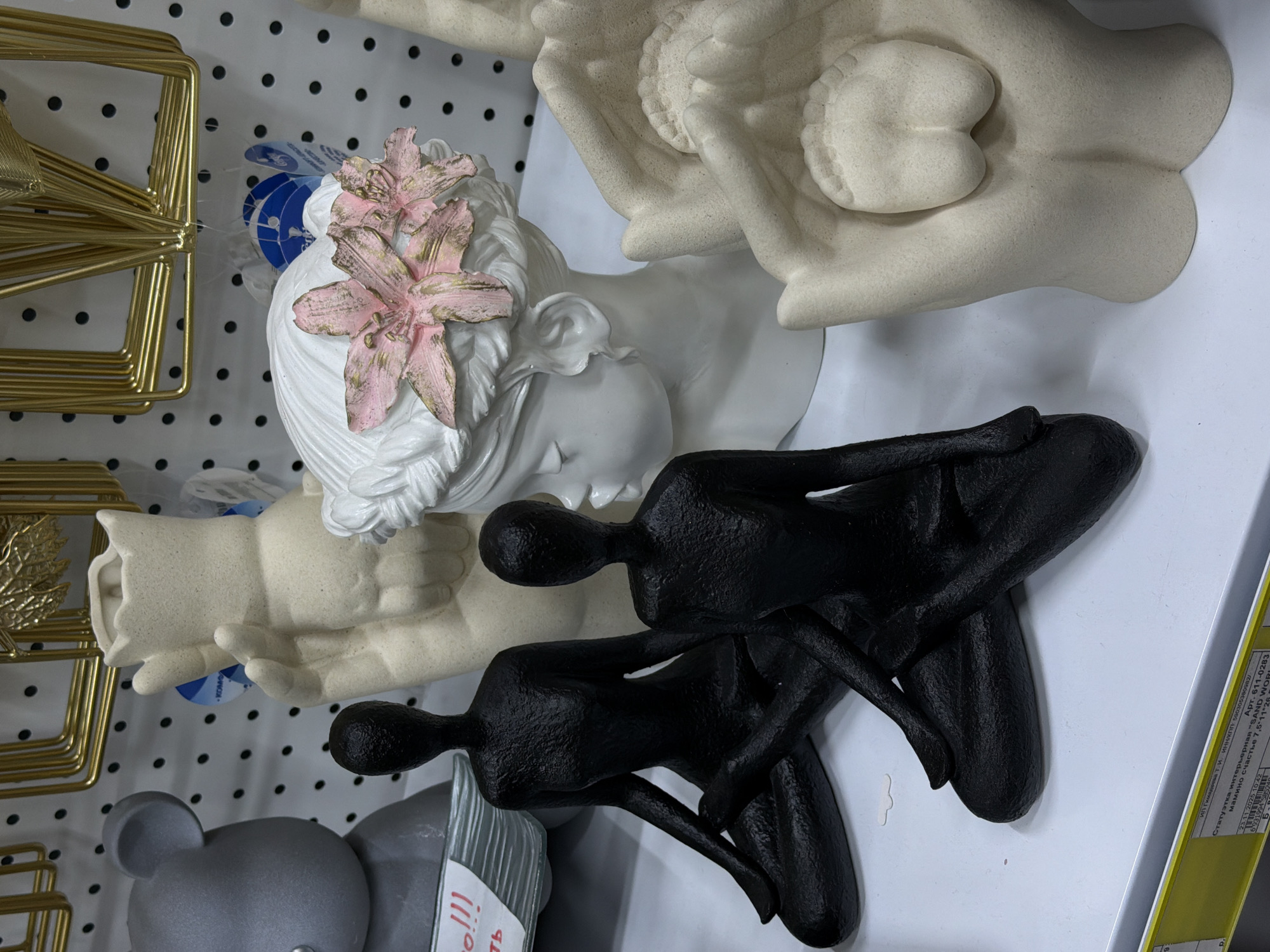 This is a still, close-up shot of a shelf in a store in Perm, Russia. The shelf is lined with decorative objects for sale. The items include:
- A sculpture of two hands holding an unborn fetus.
- A white bust of a woman with pink flowers in her hair.
- Two black figurines of a couple sitting intimately together.
- A gold metal abstract design.
- A grey bear-shaped toy.
The sticker on the shelf reads "STATUETKA INTER'ERNAYA," which means "Interior Figurine" in Russian. It also includes details of the seller, including "SAND WORLD".
