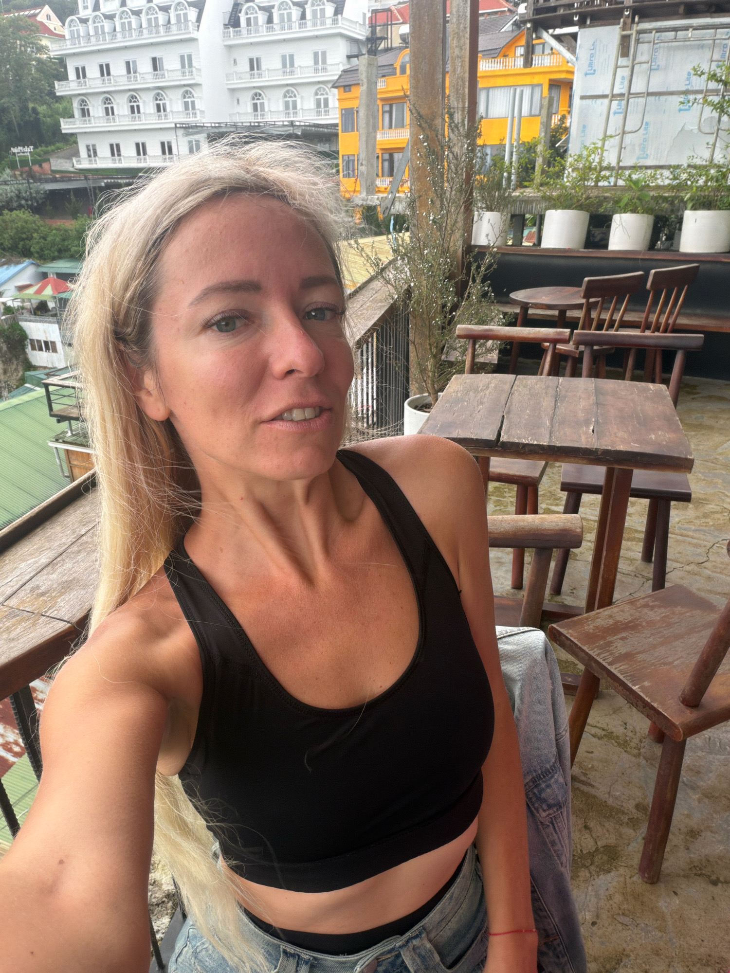 A blonde woman in a black tank top and denim shorts takes a selfie on a balcony overlooking a hillside city in Da Lat, Vietnam. The woman's face is the primary subject, with her right arm and shoulder also visible. Behind her, a balcony railing separates her from a seating area with wooden tables and chairs. In the background, multi-story buildings with white and yellow facades are nestled into the green, hilly landscape. Some of the buildings have balconies and arched windows. The sky is overcast.