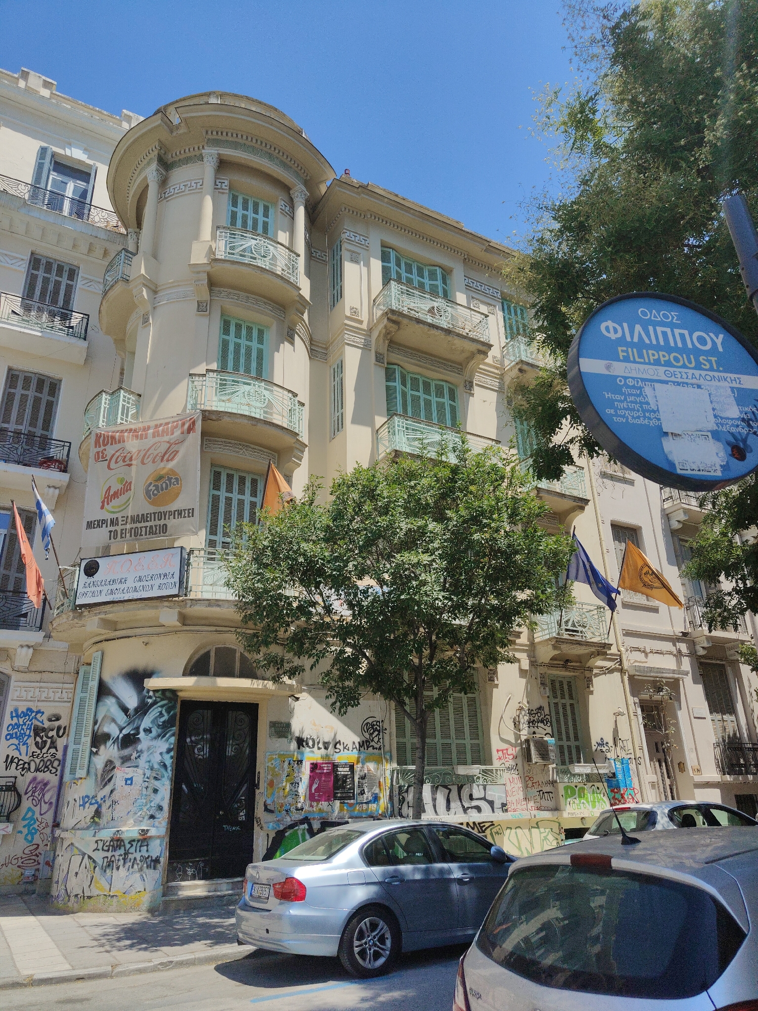 This is a full-shot, eye-level, outdoor, daytime photograph of a street scene in Thessaloniki, Greece. The focal point is a multi-story, cream-colored building with ornate architectural details and light blue shutters on the windows. The building has several balconies with decorative railings. The lower facade of the building is covered in graffiti, with a large black double door and a small arched window above it. A silver sedan is parked on the street in front of the building, facing away from the camera. A portion of another silver car is visible in the foreground, also parked on the street. A street sign with blue lettering indicating "FILIPPOU ST." and Greek text is visible on the right side of the image, partially obscured by trees. Two flags are hanging from a balcony on the building. A large green tree with dense foliage is in the middle ground, casting shadows on the building and the street. The sky is clear and blue. The overall impression is one of urban decay contrasted with the building's historical architecture.
