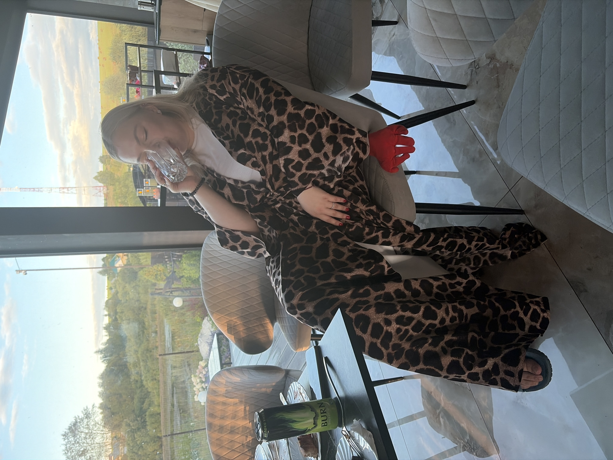 In Karsha, Russia, a blonde woman sits in a modern dining room, enjoying a drink of water from a clear glass. She is seated in a plush, light-colored chair, partially draped in a leopard-print robe that extends to the floor. Her right hand, adorned with red nail polish and a ring, rests on her leg. A bright red, glove-like object is placed on the chair next to her. In front of her, a black table holds a can of "BURN" energy drink, remnants of a meal, and a mobile phone. The room features large windows with a view of a rural landscape, including trees, a body of water, and a distant structure. The lighting suggests it is late afternoon or early evening.