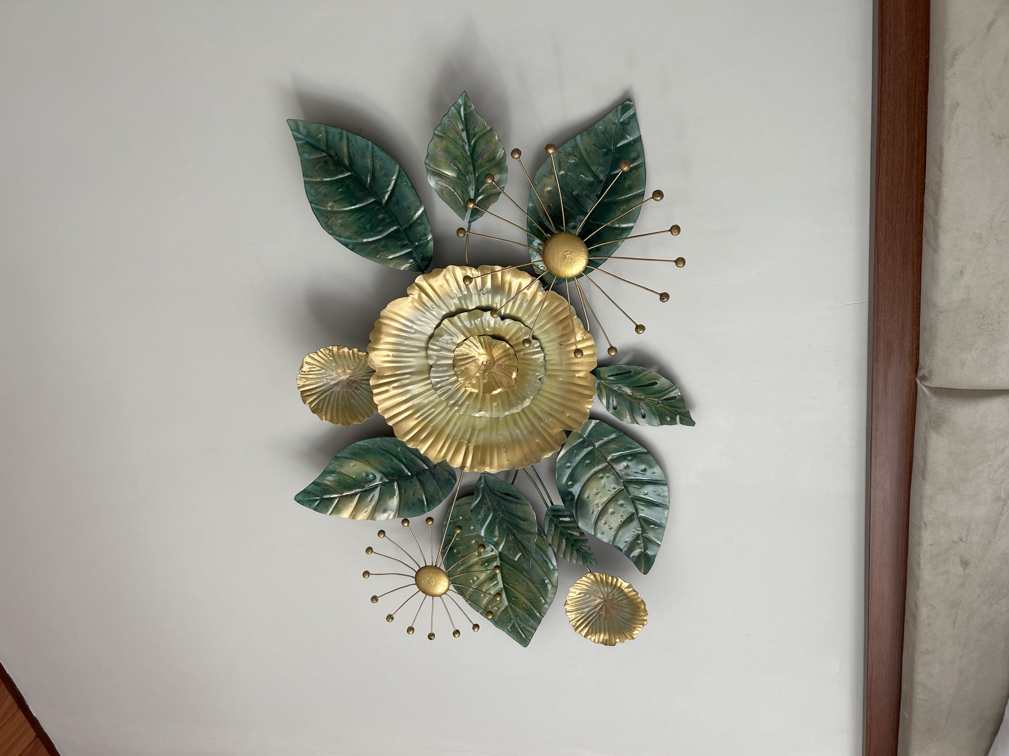 A close-up, eye-level shot captures a metallic wall art sculpture mounted on a plain, light-colored wall in Abiansemal, Indonesia. The sculpture depicts a stylized floral arrangement with large, metallic green leaves and golden flower elements. The central flower is large and golden, with multiple layered petals. Smaller golden circular elements, some resembling unfurling buds and others with radiating thin metal rods tipped with small golden balls, are interspersed among the leaves. The leaves themselves are detailed with vein patterns and have a slightly textured, metallic sheen. The overall impression is one of artistic craftsmanship, possibly a decorative piece for a bedroom or living area, given the headboard visible at the bottom of the frame. The lighting appears to be natural, casting subtle shadows that give the sculpture depth.