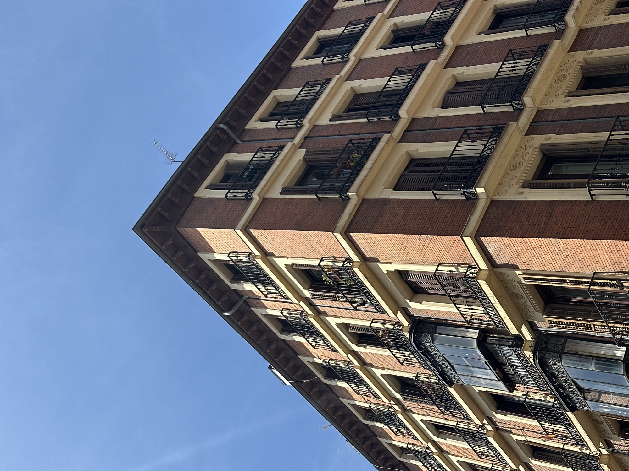 This image captures a low-angle, tilted view of a multi-story building against a clear blue sky, set in Madrid, Spain. The building is constructed primarily of reddish-brown brickwork, punctuated by regularly spaced windows. Each window is framed by light-colored, likely cream or beige, architectural elements and features dark wooden shutters.
A prominent detail across the facade is the ornate black wrought-iron work, appearing as either full balconies or decorative window guards, adding a classic European architectural touch. Some of these ironworks exhibit elaborate scroll patterns. Detailed decorative carvings, including floral or geometric motifs, are visible on the light-colored sections surrounding some windows, particularly in the upper stories.
The top-left edge of the building's roofline is visible, with a dark gutter pipe running along it. A small, silver television antenna protrudes from the rooftop, silhouetted against the bright sky. The dramatic angle of the photograph emphasizes the building's height and the regularity of its architectural design. The lighting suggests it is daytime, with clear, sunny weather. No people or visible text are present in the image.