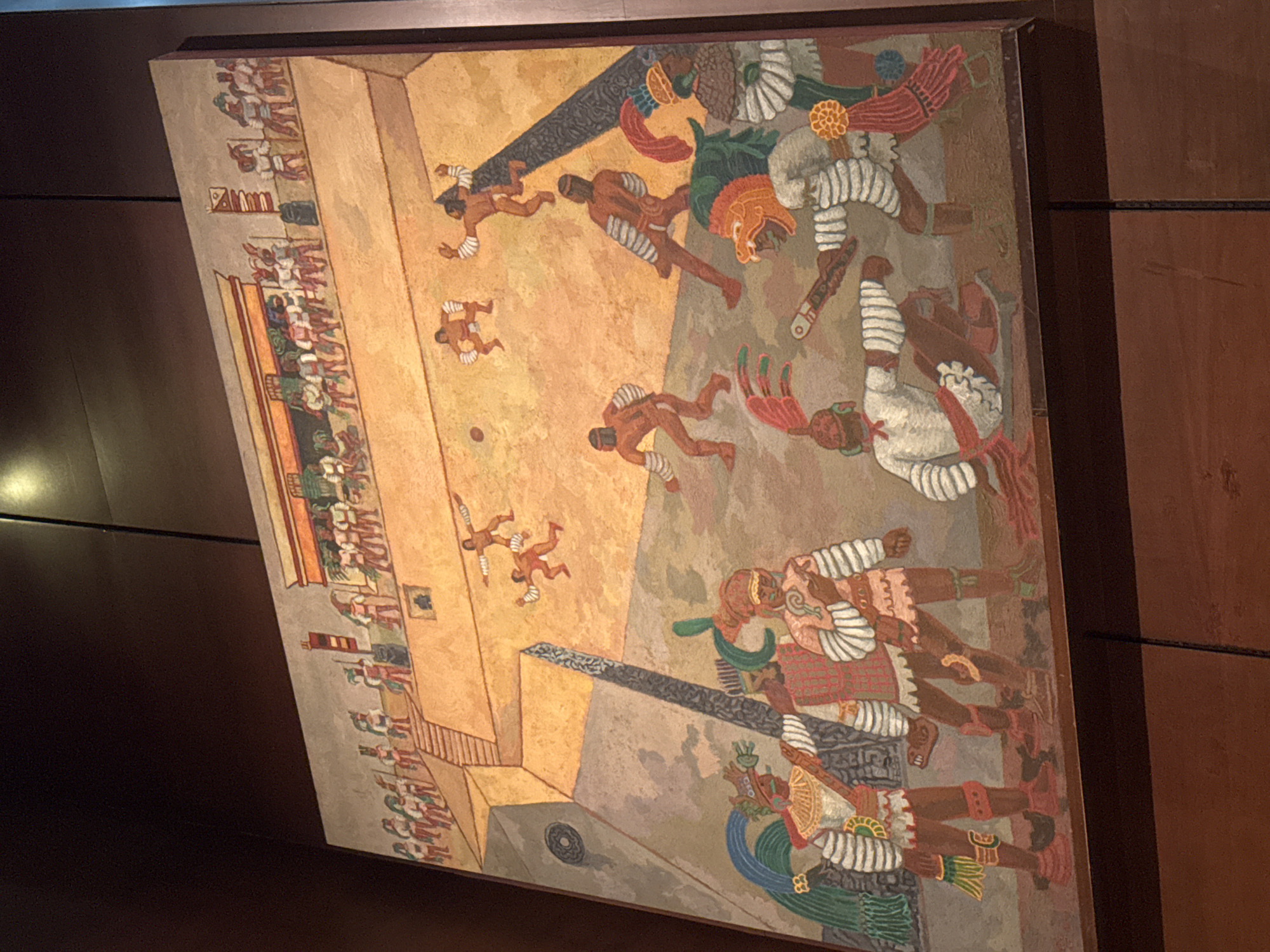 The image depicts a mural in Mexico City, Mexico, portraying an Aztec ball game. The mural shows players in loincloths with protective gear, maneuvering to strike a ball. Spectators are seated along the side of the court. The artwork is done in a stylized, illustrative manner, resembling pre-Columbian art. The background is a combination of warm earth tones and cooler grays. The mural is framed, mounted on a wall with brown wood panels.