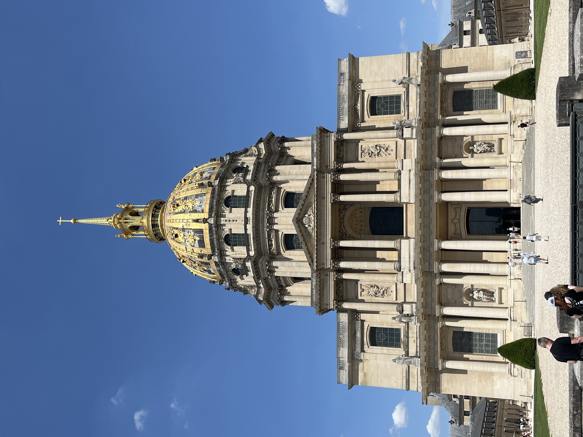 This image shows the Hôtel des Invalides in Paris, France, under a clear blue sky with scattered clouds. The grand building features a prominent golden dome topped with a spire and cross. The facade is made of stone and adorned with classical columns and sculptures. Several people are walking on the gravel pathway leading up to the entrance, some are in groups, others are walking individually. Two conical trees are situated on either side of the entrance steps. The surrounding area appears to be a well-maintained plaza or garden.
