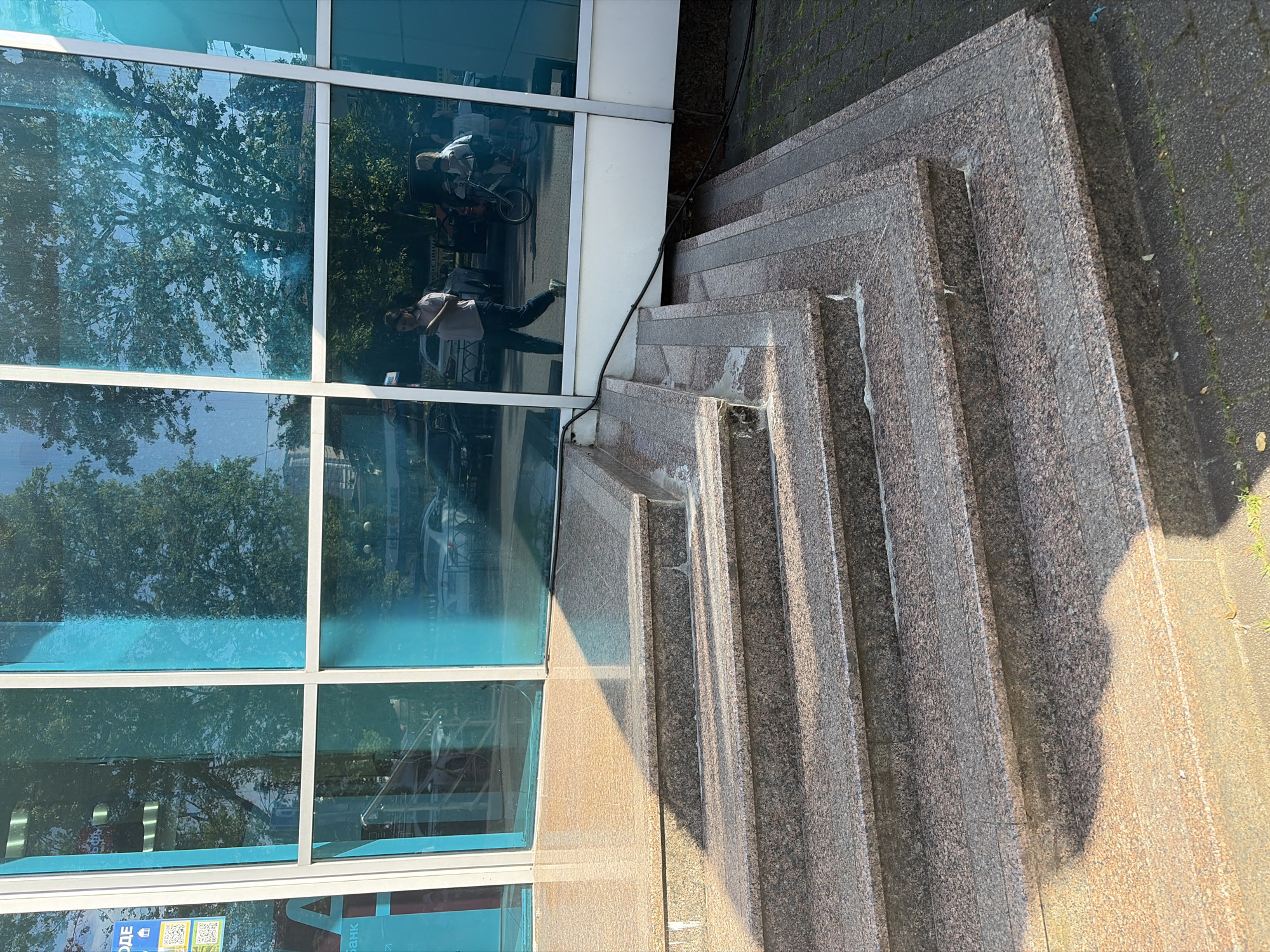 The image shows a close-up view of a modern building entrance with a granite staircase and large blue glass windows. The windows reflect trees and people outside. In the reflection, a woman is walking, and a person is on a bicycle. The staircase is well-maintained, with visible grout lines between the granite slabs. A black cable runs across the bottom of the image. The building appears to be located in an urban area.