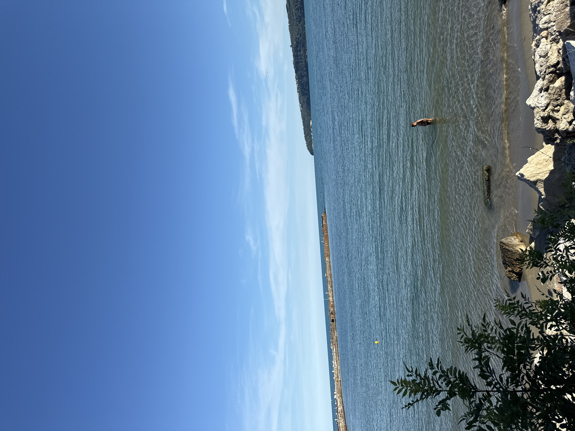 A person is swimming in a calm, blue body of water. The water is clear enough to see the sandy bottom near the shore. The person is in the middle distance, facing away from the camera, with their head and shoulders above the water. To the right of the person, there is a rocky shoreline with some vegetation growing on top. In the background, a long, narrow pier or breakwater extends into the water. A yellow buoy is visible in the water to the left of the pier. The sky is a clear, bright blue with a few wispy white clouds. The sun appears to be high in the sky, suggesting it is daytime. The overall scene conveys a sense of peace and relaxation, with the person enjoying a swim on a sunny day.