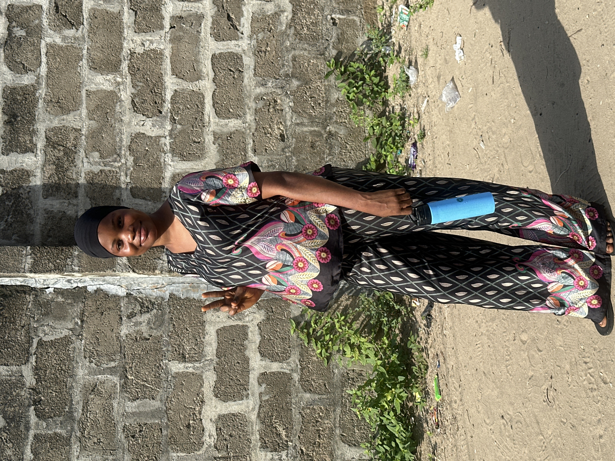 A young woman stands outdoors, smiling and giving a peace sign with her right hand. She is wearing a dark headscarf and a two-piece outfit consisting of a black top and matching pants, both adorned with a vibrant floral and abstract pattern featuring pink, orange, green, and white elements. Her left hand holds a light blue cylindrical bottle.
The setting features a textured, grey concrete block wall behind her, showing signs of wear and tear. The ground she stands on is sandy or dirt-covered, with patches of green weeds and scattered white plastic litter. Strong sunlight casts distinct shadows, suggesting a bright and sunny day, possibly mid-day or early afternoon. The woman appears relaxed and friendly. A partially visible word "YOUNG" can be seen in white text on her top, near her right shoulder. The environment gives the impression of a casual outdoor space, likely in an urban or semi-urban area in Badagry, Nigeria.