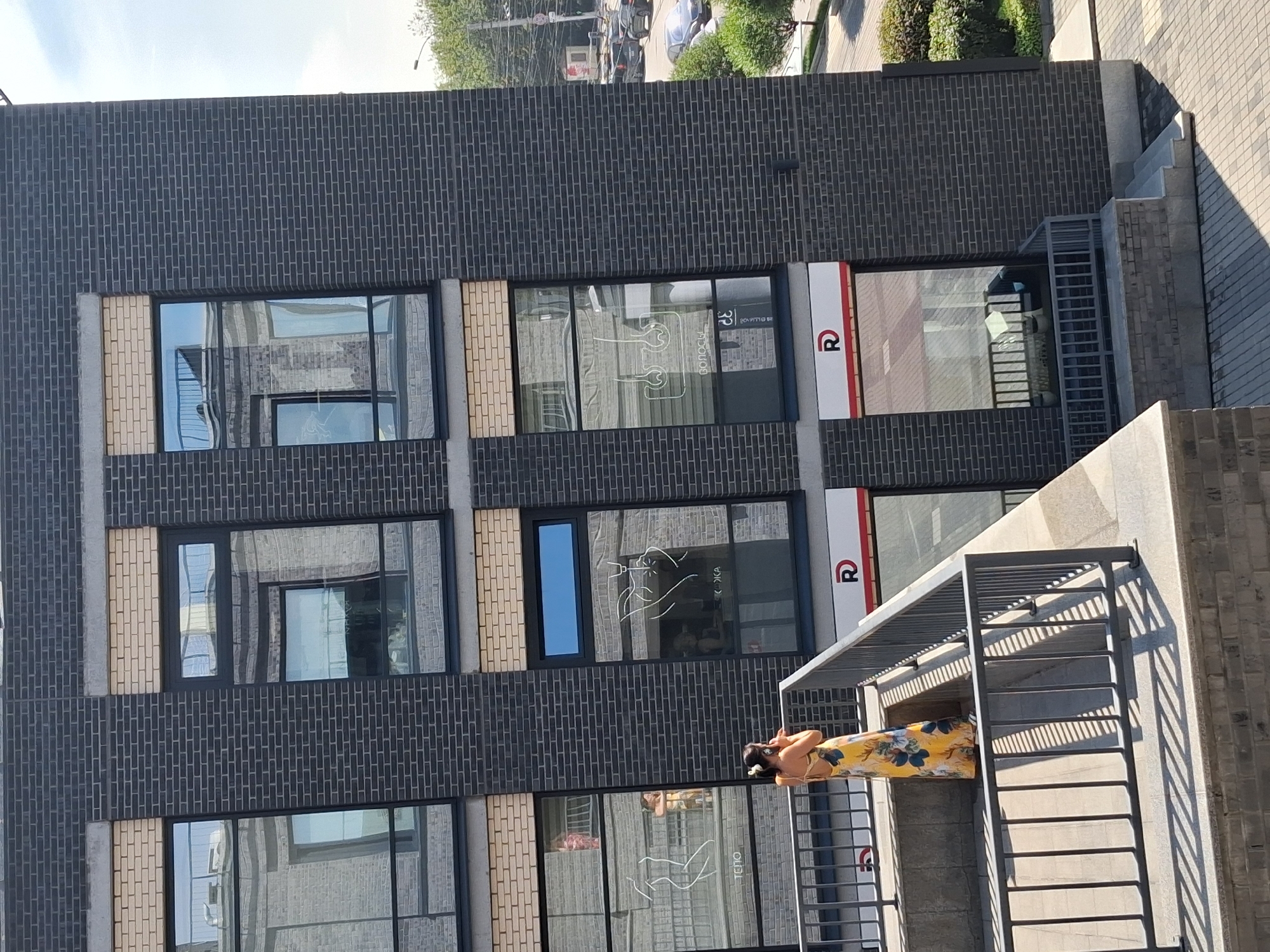 The image depicts a woman standing on a balcony on a sunny day, wearing a yellow floral dress and headphones. She is looking towards her right, away from the camera, and appears to be holding a phone to her ear. The balcony overlooks a street with a modern building featuring dark brick and large windows. Inside the windows, there are graphics related to beauty services, including outlines of a human body and hair follicles. The building has signage with the letter "R" in a red circle. In the background, cars are visible on the street, and trees line the sidewalk. The scene appears to be in an urban environment. The provided information states the location is in Novosibirsk, Russia.