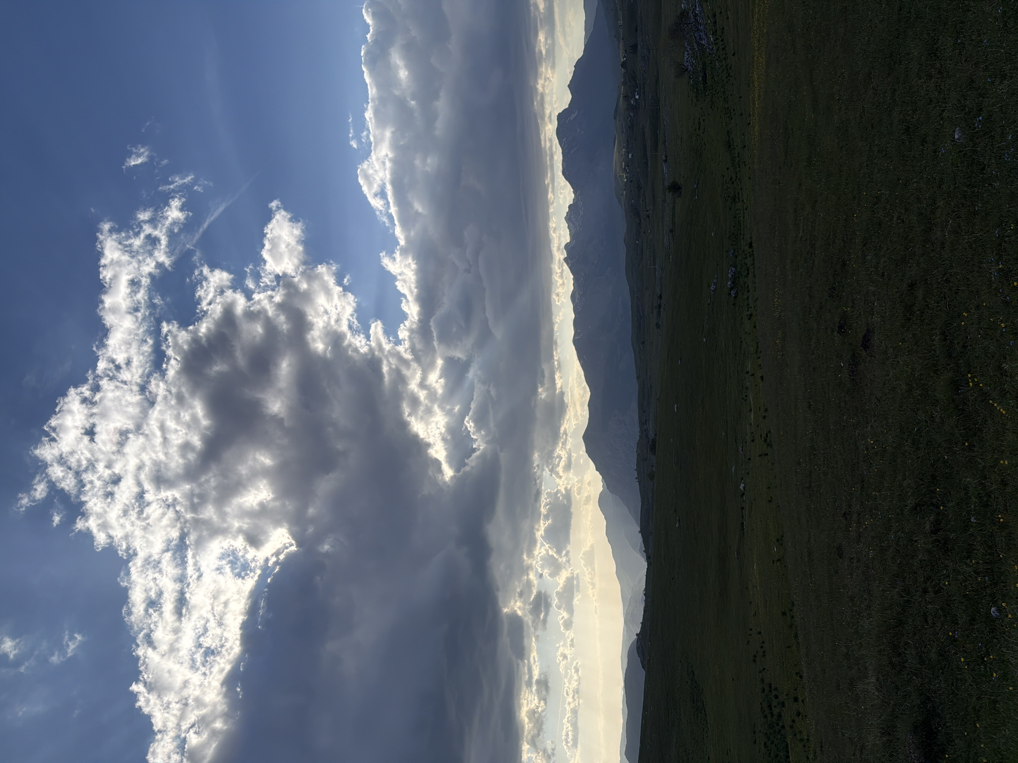 The image shows a landscape in Pišče, Montenegro. The sky is partially covered with clouds, some of which are backlit by the sun. Mountains are visible in the midground, fading into the distance with a light haze. A grassy field occupies the foreground.