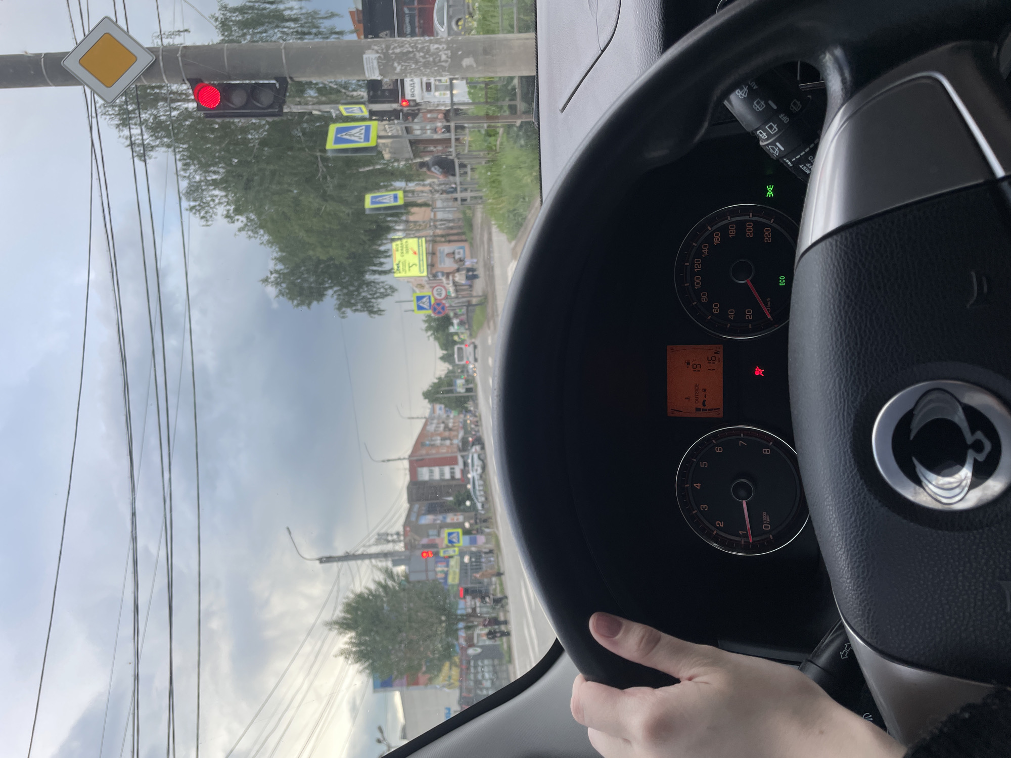 The image, taken from the driver's perspective inside a car, captures a street scene in Votkinsk, Russia, with the vehicle stopped at a red traffic light. The sky is overcast with clouds, indicating a cloudy daytime setting.
In the foreground, a person's hand rests on the bottom of a black steering wheel, which features a silver trim and a prominent logo in the center. The car's dashboard is partially visible, showing a speedometer indicating 0 km/h and a tachometer also at 0 RPM, confirming the car is stationary. A digital display shows "OUTSIDE 19," likely indicating an outdoor temperature of 19 degrees Celsius, and the number "115." Illuminated indicator lights include a red traffic light icon, a red seatbelt warning, and a green headlight symbol. A wiper control stalk, with "MIST" visible, is positioned to the right of the steering wheel.
Beyond the windshield, a red traffic light is prominently displayed, confirming the stop. Above it, a white square sign with a yellow diamond, indicating a main road, is attached to an overhead structure. Numerous utility poles and wires crisscross the sky. Along the street, multiple blue rectangular signs with white pedestrian crossing symbols are visible. Buildings, some of which are multi-story and appear residential, line the road in the distance. Other vehicles and pedestrians can be seen further down the street. Various road signs with Cyrillic text are present, including one with "40" in a red circle (a speed limit sign) and partially visible text "ВОДА" (water) on another sign.