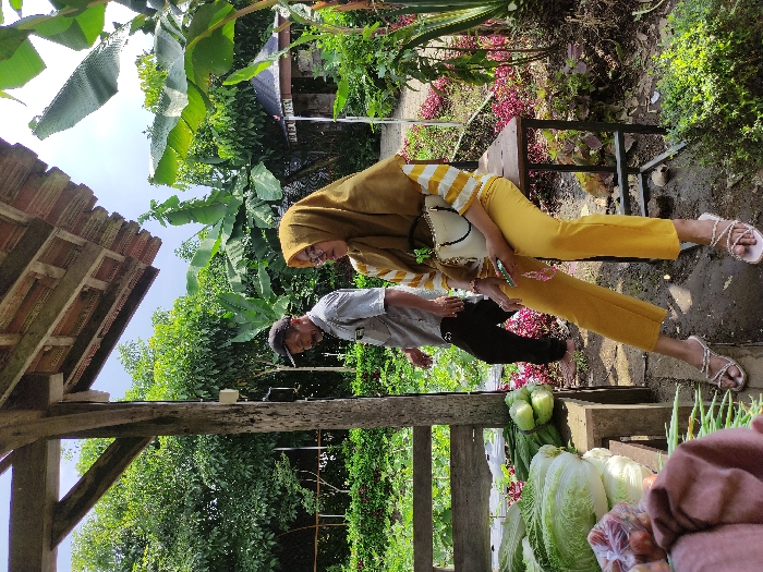 The image shows a woman sitting on a wooden stool with a man standing behind her in the background. The woman is wearing a yellow headscarf with a striped white and brown blouse and yellow pants. She is holding a white handbag. The man is wearing a dark blue shirt and jeans, and a dark blue cap. They are standing near a wooden stall with produce for sale. The woman is looking to the left, and the man is looking down. The image is taken in a market in Kecamatan Pujon, Indonesia. The image is taken outdoors, and the background shows vegetation.