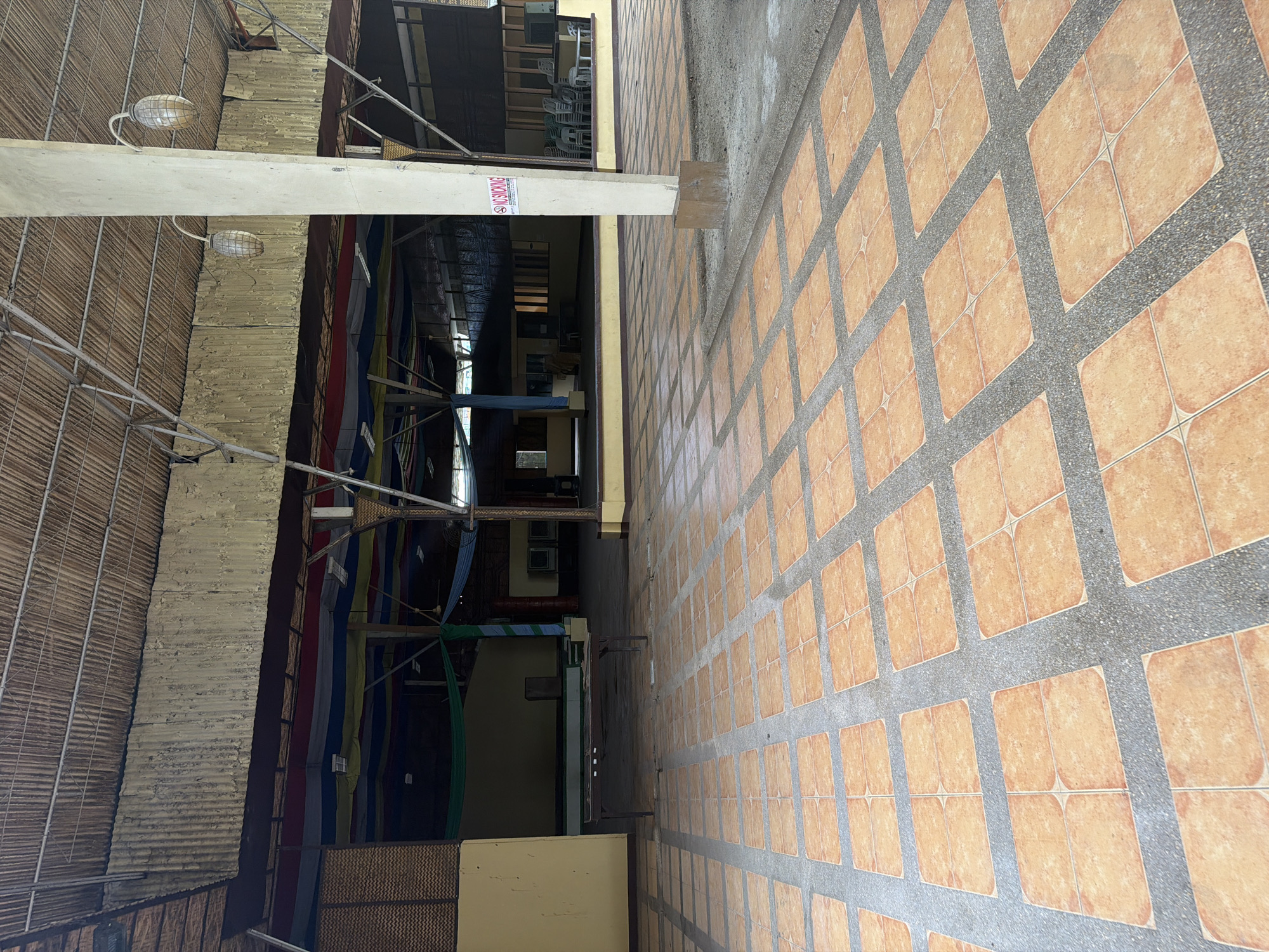 The image shows an outdoor space in what appears to be Davao City, Philippines. The area is covered with orange square tiles separated by gray concrete lines. To the left of the tiled area is a covered walkway or hallway, possibly part of a commercial building or market. The ceiling of this walkway is partially made of corrugated metal or woven material, with visible support beams and light fixtures. A concrete pillar supports the structure, and a "NO SMOKING" sign is attached to it. The space is dimly lit, and the interior appears to contain tables and chairs. Colorful cloth or banners hang along the sides of the walkway.