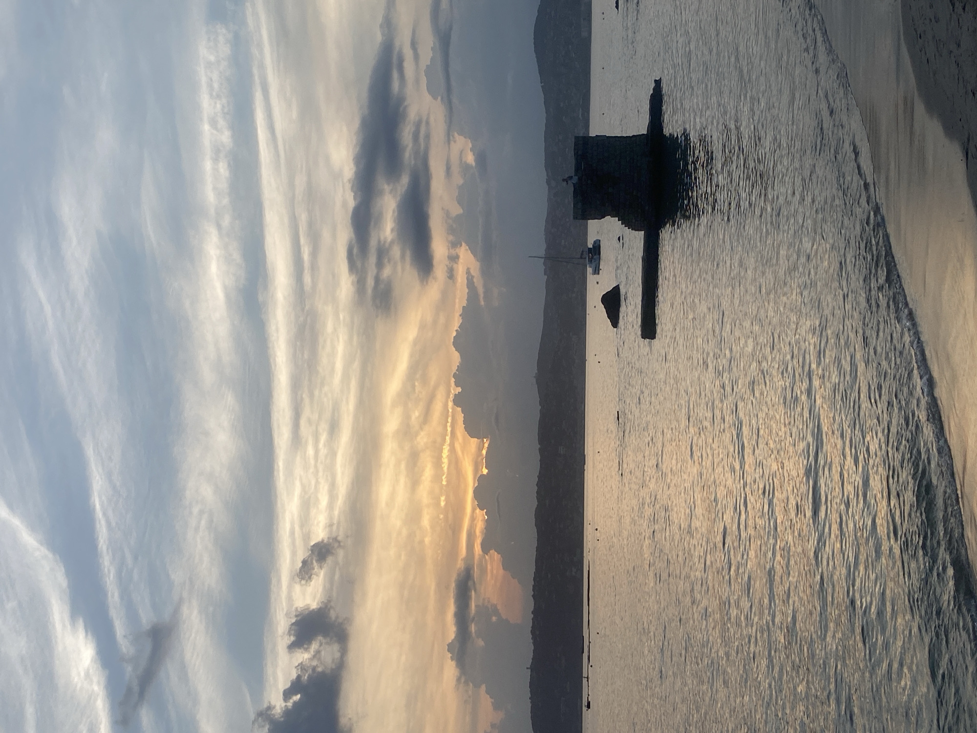 The image depicts a serene coastal scene at sunset in Antibes, France. The sky is filled with a dramatic display of clouds, transitioning from pale blue at the top to warm hues of orange and yellow near the horizon, suggesting the sun is setting or has recently set. Wispy, thin clouds streak across the upper sky, contrasting with darker, more voluminous clouds gathering over the distant coastline.
In the foreground, the calm sea reflects the soft light of the sky, creating shimmering patterns on its surface. Gentle waves lap against a sandy beach, visible at the bottom right of the frame, with the sand appearing damp and reflecting the muted light.
Further out in the water, a small, weathered stone tower stands on a low, rectangular platform, partially submerged. A solitary figure stands atop the tower, silhouetted against the sky, appearing to be observing the sunset. To the left of the tower, a large, dark rock is visible, and beyond that, several buoys or small markers are scattered across the water.
In the middle distance, a sailboat with its mast prominently displayed glides across the water, its white sails catching the light. The distant shoreline is lined with hills and vegetation, with some structures or lights faintly visible along the coast, indicating a populated area. The overall atmosphere is one of tranquility and natural beauty.