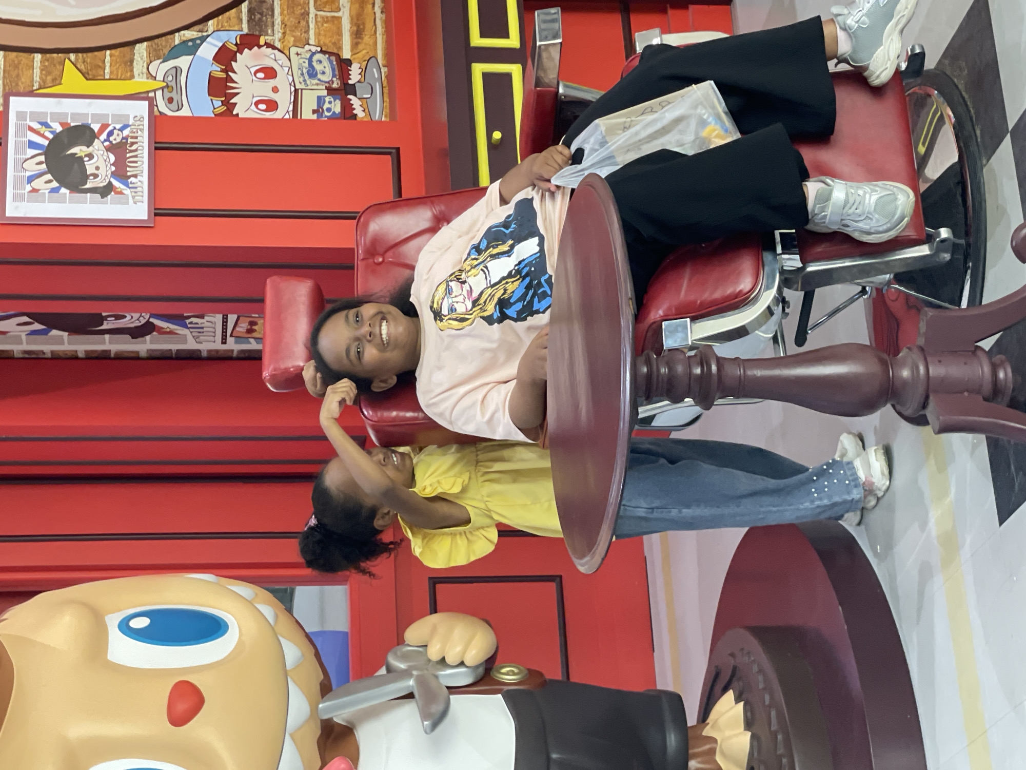 In Pattaya City, Thailand, two young girls are posed in a stylized barbershop setting. One girl, on the left, wears a yellow ruffled dress and blue jeans, her arm extended towards the other girl. The second girl, seated in a red barber chair, is wearing a pale pink t-shirt with a graphic of a blonde woman and black pants. She is smiling and holding a clear plastic bag. Behind the girls, red vertical panels and framed cartoon artwork adorn the wall, contributing to the playful, commercial atmosphere. A large cartoon figurine, resembling a character from a Japanese toy line, stands to the left of the frame, its large blue eyes and open mouth a prominent feature. A dark red, round table is positioned in the foreground, obscuring the lower portion of the scene. The flooring consists of white tiles with yellow and black stripes.