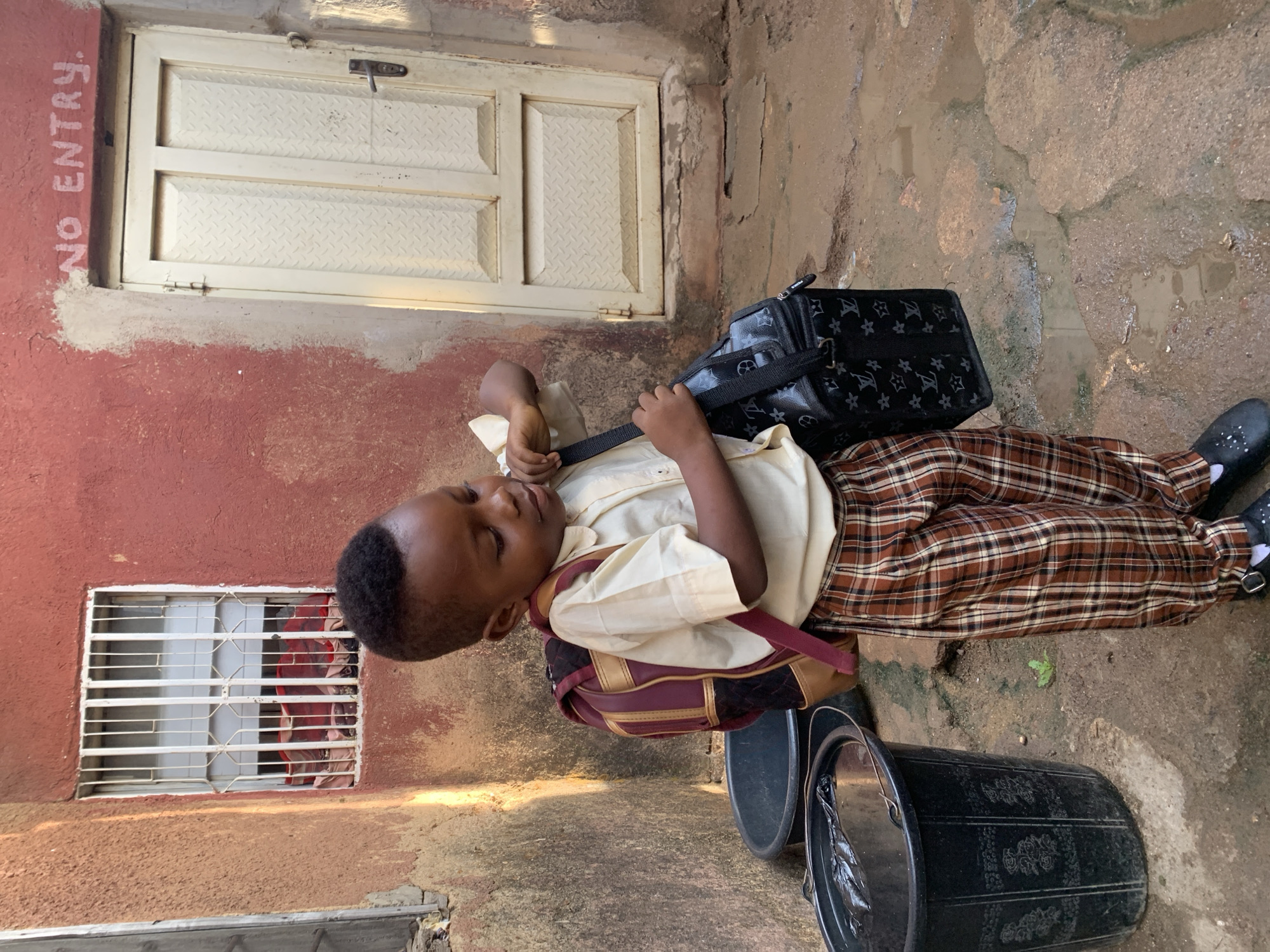 The image shows a boy in a school uniform, wearing a light yellow shirt, plaid brown pants, and black shoes with white socks. He has a black bag with a patterned design slung over his shoulder. The environment appears to be an outdoor area in Jos, Nigeria. There is a building with a red-painted wall and a white metal door with a "NO ENTRY" sign written on it. Another window with white bars is visible on the same wall. On the ground, there are two black buckets with ornate designs. The ground is a mixture of dirt and concrete with some wet spots. The overall setting suggests a residential or commercial area with signs of wear and tear.