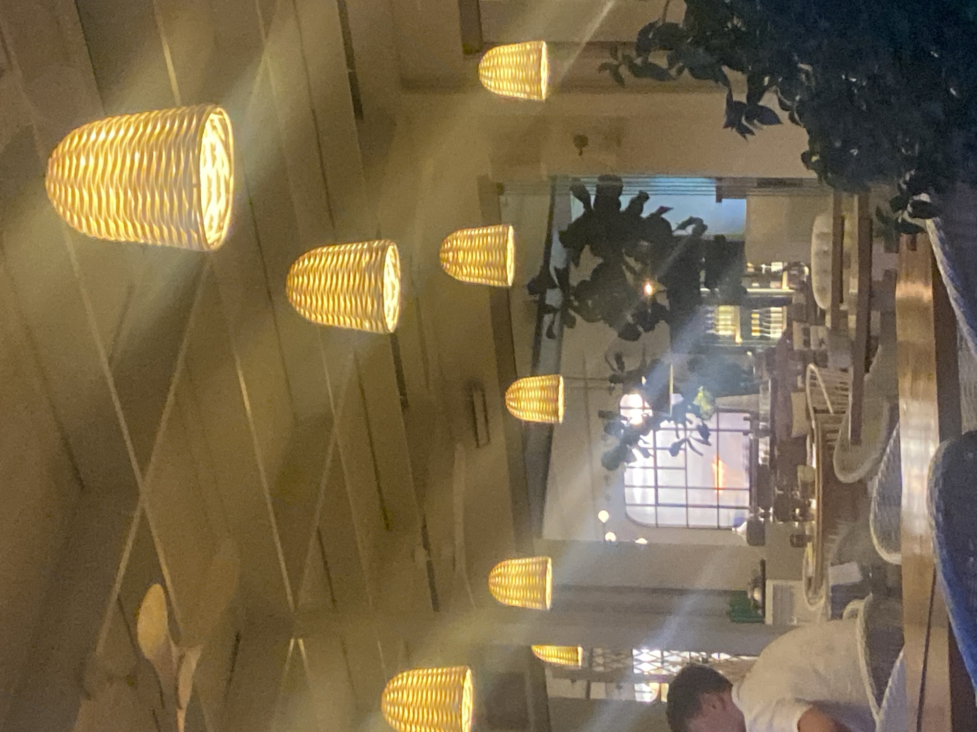The image depicts the interior of a restaurant, likely in Estepona, Spain, during what appears to be twilight or evening hours, given the warm glow of the lighting. Several woven pendant lamps, shaped like elongated baskets, hang from a light-colored, slatted ceiling. These lamps cast a warm, yellow light that illuminates the space.
In the foreground, a person is partially visible on the left, wearing a white shirt and appearing to be seated at a table. The midground reveals tables and chairs, suggesting a dining area. Some tables have placemats and what appear to be place settings. Through a large window in the background, a sunset is visible, with hues of orange and pink painting the sky. The window has a grid pattern, reminiscent of classic architecture. Lush green foliage is present, both inside the restaurant and potentially outside, adding a touch of nature to the scene. The overall ambiance is warm and inviting, with a clear focus on the lighting and the view through the window.