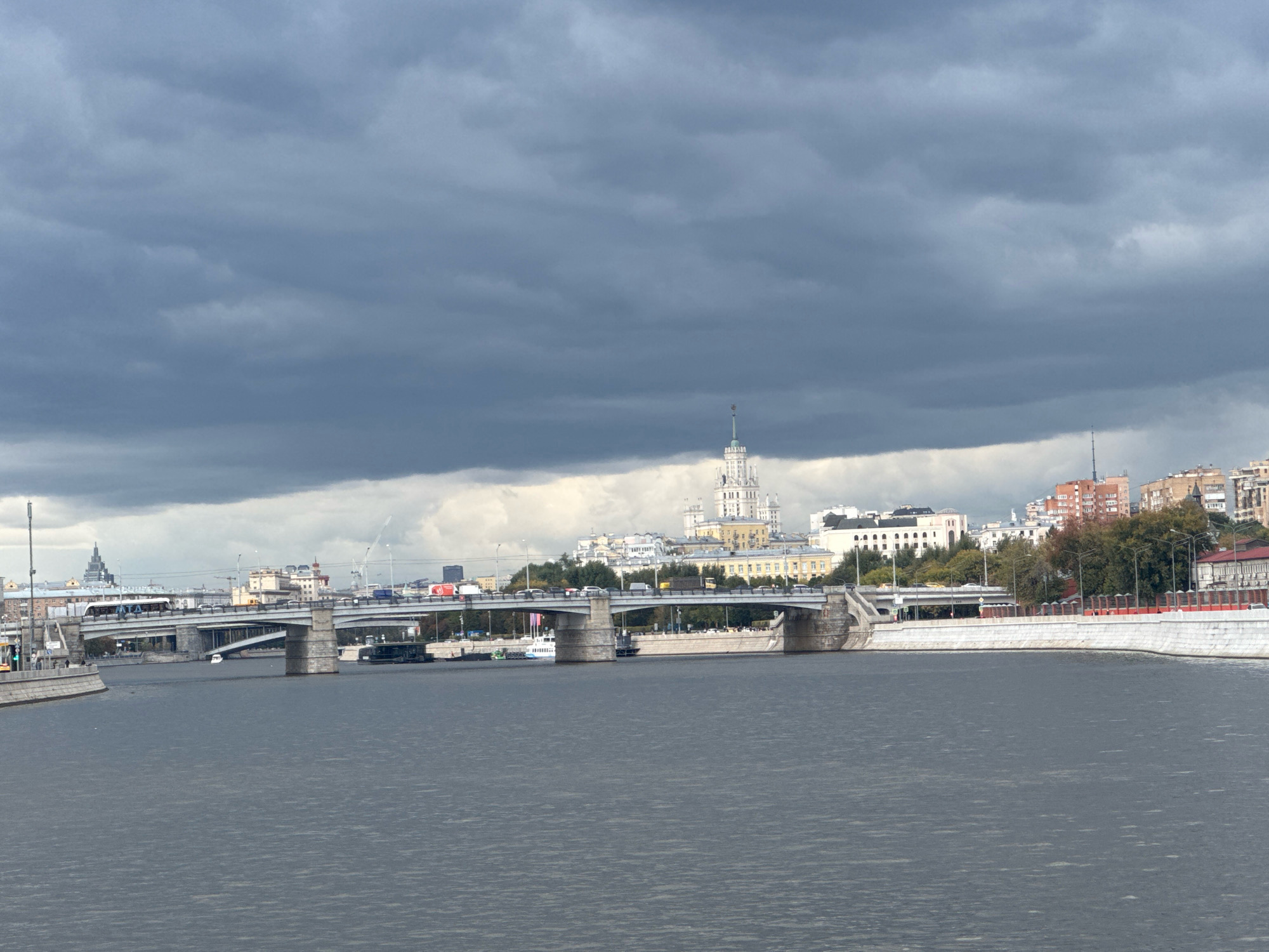 This is an image of the Moscow River in Moscow, Russia, with a view of the Stalinist skyscrapers in the background, specifically the Moscow State University (MGU) main building, which is prominently featured. The sky is overcast with dark, heavy clouds. A wide bridge, likely carrying vehicular and pedestrian traffic, spans the river in the foreground. Several boats or barges are visible on the water, and the riverbanks are lined with stone embankments and buildings. The overall atmosphere is somewhat somber due to the cloudy weather.