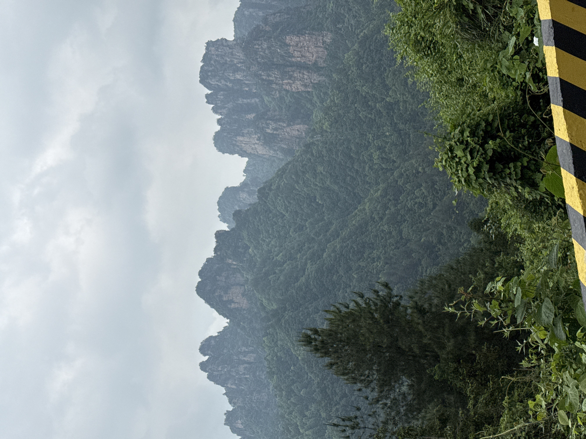 The image shows a mountainous landscape in Zhonghu, China under an overcast sky. The mountains are covered in dense, green vegetation. A black and yellow striped barrier stands to the right. The foreground features a mix of trees and shrubs, while the background shows the rocky peaks of the mountains. The light is diffused due to the cloudy weather.