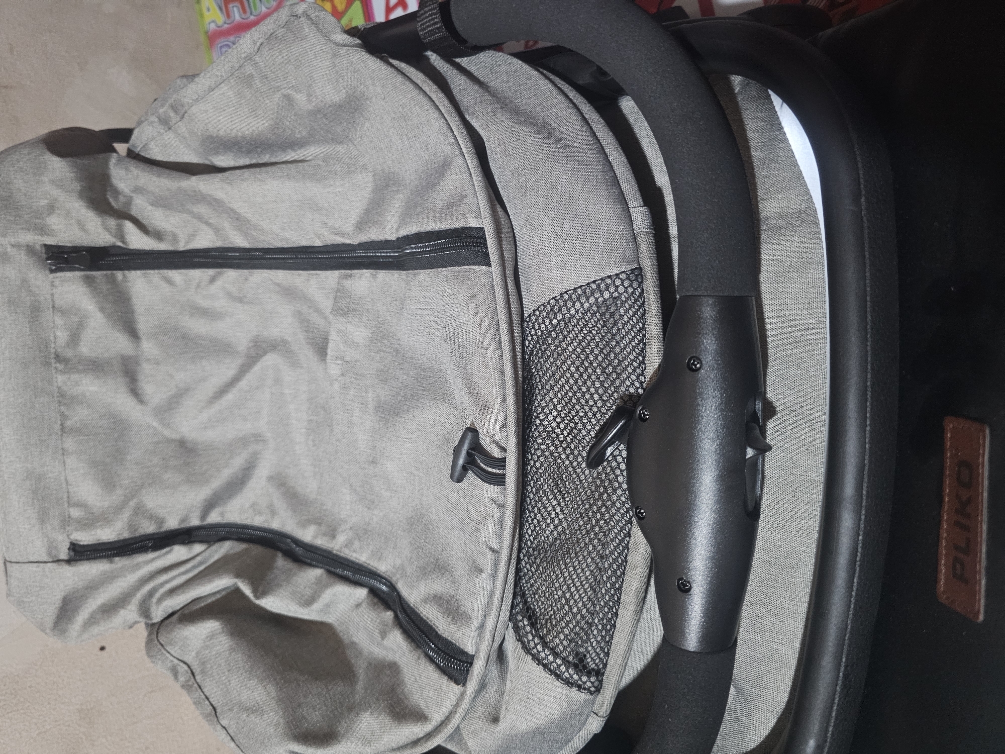 The image shows a close-up view of a grey baby stroller. The stroller has a black zipper running vertically down the center of the main fabric. A black drawstring closure with a plastic toggle is visible below the zipper. There is a mesh panel on the lower part of the stroller. The stroller's handle is black and appears to be padded. A small brown leather tag with the word "PLIKO" embossed on it is attached to the lower section of the stroller. The background is a light-colored, textured wall, suggesting an indoor setting. There are no people visible in the image. The location is the city of Dadap, Indonesia.