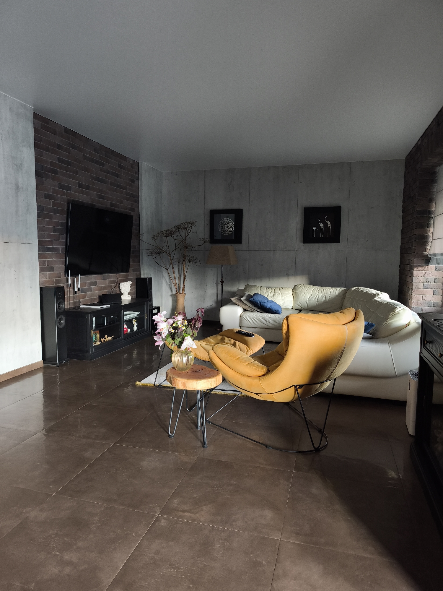 The image depicts a modern living room with an industrial-chic aesthetic. The walls are a mix of exposed dark brick and textured concrete panels. A large flat-screen television is mounted on the brick wall above a black media console. To the right of the television are two black speakers. In the center of the room, a plush, mustard-yellow armchair with a black metal frame is positioned in front of a large, white leather sofa. A small, round wooden side table with hairpin legs sits in front of the armchair, holding a vase of pink and white flowers and a dried decorative branch. The floor is covered in large, dark brown tiles with a glossy finish, reflecting the light. Two framed pieces of art depicting stylized birds are hung on the concrete wall behind the sofa. A floor lamp with a tan shade is visible to the left of the sofa. The room is bathed in natural light, casting dramatic shadows from the furniture. The scene appears to be a still, unpopulated interior space. It is not possible to determine the specific location within the city of Kaluga, Russia, from the visual information provided.