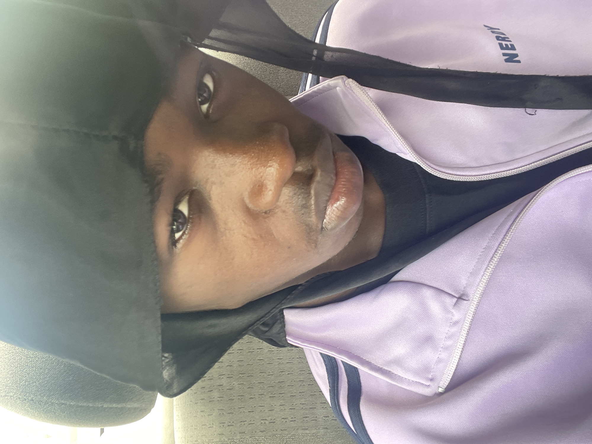 A portrait of a young Black man wearing a black durag and a lavender-colored tracksuit jacket with navy blue stripes. The brand "NERDY" is partially visible on the jacket. The background appears to be an indoor setting, possibly a car, with neutral-colored upholstery or seats. No specific details are visible that would identify the location as Ado, Nigeria. The focus is on the man's face, captured in soft, natural lighting.