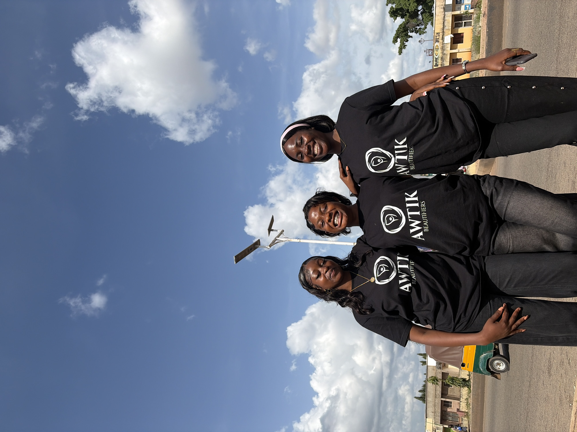 Three young women are standing outdoors in what appears to be a city street in Gura Top, Nigeria. They are all wearing matching black t-shirts with a white logo that says "AWTİK BEAUTIFIERS". The woman on the left has long, dark, wavy hair and is wearing a gold necklace. The woman in the middle has dark, short hair and is smiling broadly, showing her teeth. The woman on the right has dark, short hair and is wearing a pink headband. She is also smiling. Their arms are around each other. In the background, there is a blue sky with white clouds. There is also a street lamp, a yellow and green auto-rickshaw, and some buildings. The women are standing on a paved surface, likely a sidewalk or road. The overall impression is a casual, outdoor gathering.