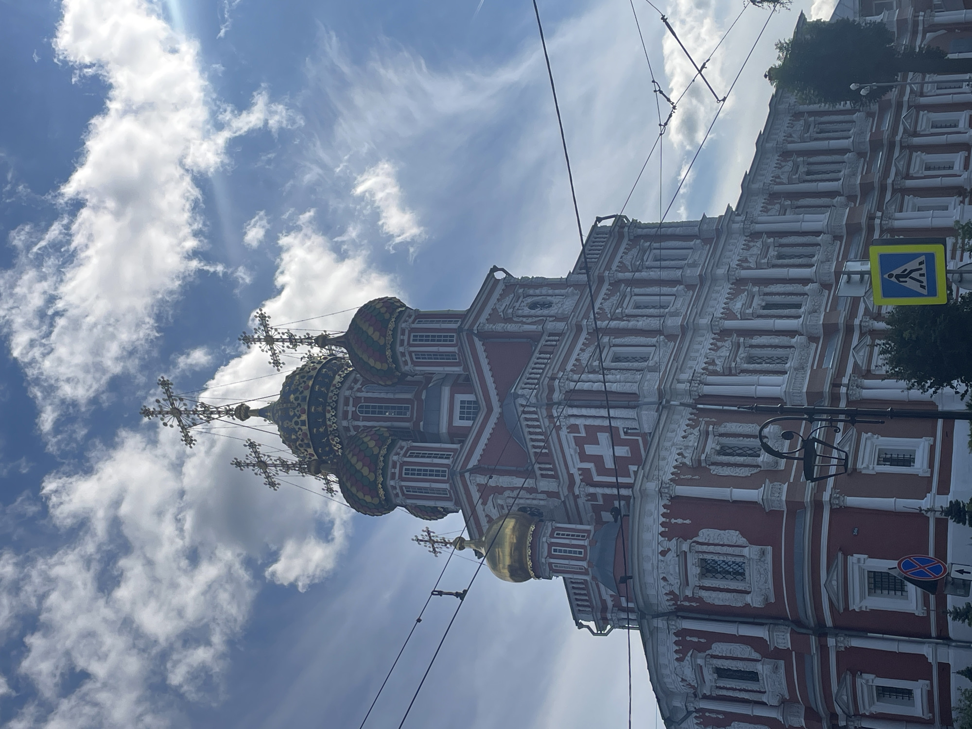 The image shows a view of the Church of the Nativity of the Virgin Mary in Nizhny Novgorod, Russia. The church is a red and white building with multiple domes, the largest of which is gilded. It is a sunny day with a blue sky and white clouds. There are overhead wires in the foreground, suggesting the presence of a tram or trolleybus line. A pedestrian crossing sign is visible on the lower left of the image, and a street lamp is positioned near the right side of the church. The architecture appears ornate, with intricate detailing on the facade.