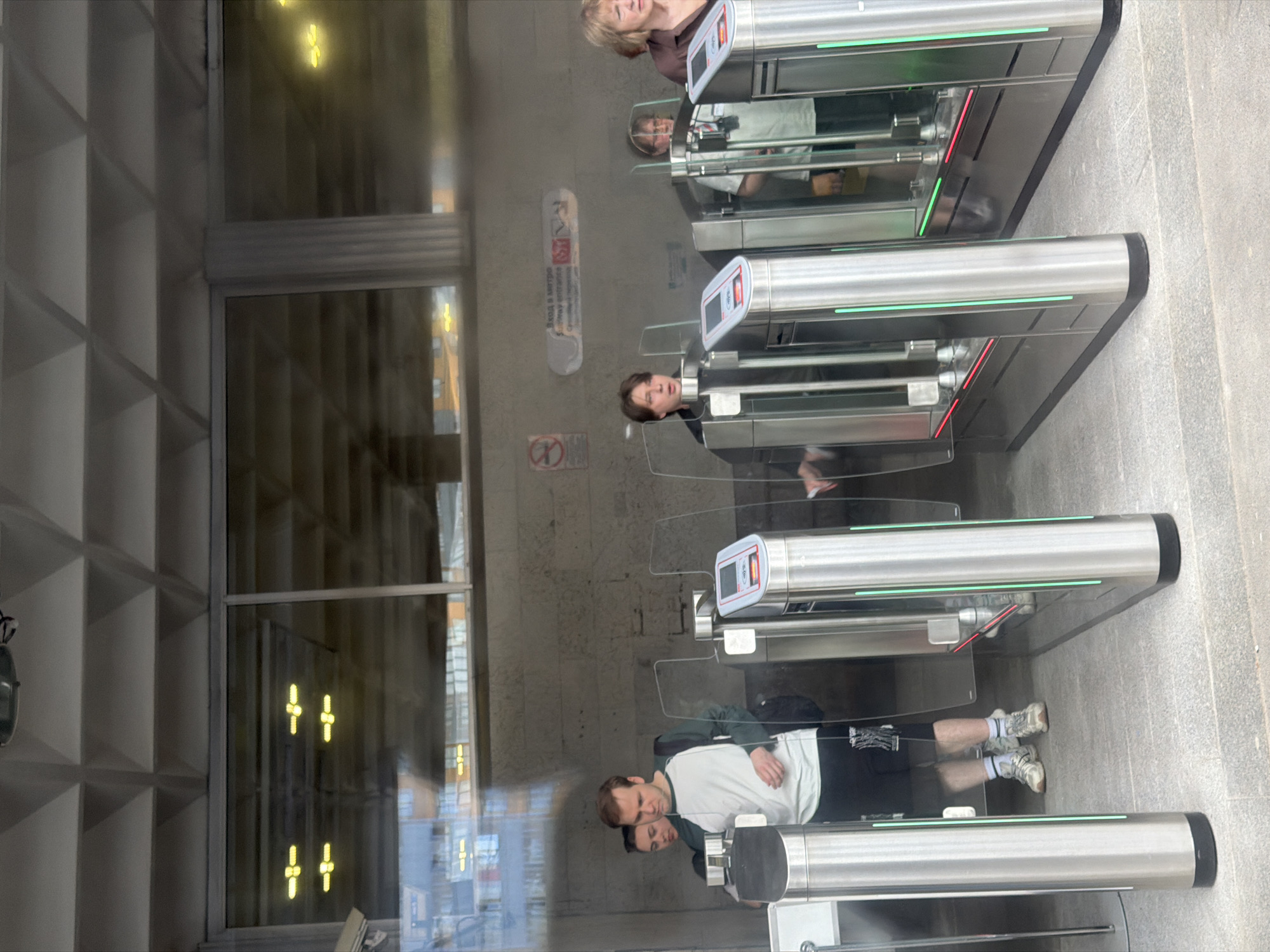 The image captures a view of individuals passing through a turnstile system, likely at a subway entrance, in an urban environment. The turnstiles are modern, constructed of stainless steel with illuminated green and red indicator lights, suggesting a functional entry point. Several people are visible in various stages of passage, with one man in the foreground appearing to interact with the turnstile. Behind them, a sign with Cyrillic text and pictograms is mounted on a concrete wall, with the partial text "вход в метро" which translates to "subway entrance" visible. The background reveals reflections in a large glass pane, showing an interior space with rows of yellow lights and the ceiling structure of the building, which appears to be a modern architectural design with geometric patterns. The overall scene suggests a typical day at a public transportation hub, with the urban setting implied by the infrastructure and the presence of commuters. The specific location is identified as Unknown, Russia.