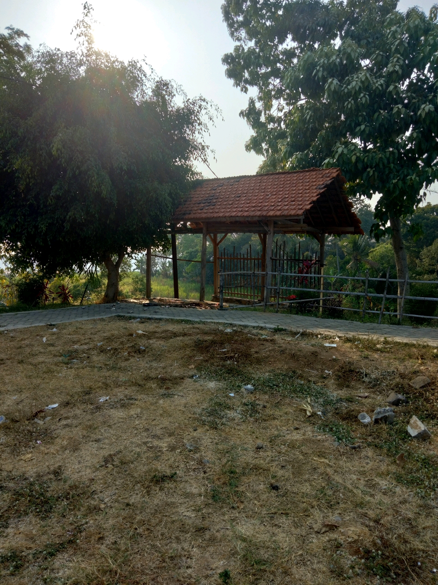 The image depicts an outdoor scene in the city of Sawahan, Indonesia, bathed in the warm light of what appears to be either early morning or late afternoon sun. In the foreground, dry, yellowish-brown grass covers the ground, interspersed with small patches of green vegetation and scattered debris, including white plastic wrappers and small stones. A paved walkway, constructed from interlocking gray pavers, leads from the lower left towards the center of the frame.
Dominating the midground is a small, open-sided wooden structure with a tiled, terracotta-colored roof. This structure appears to be a shelter or pavilion. It is supported by wooden posts and features a rustic fence made of horizontal wooden poles. Behind this structure, a dense arrangement of trees and vegetation forms the backdrop, with lush green foliage and a few palm trees visible.
A large, mature tree with dark green leaves stands prominently to the left of the shelter, its branches extending towards the sun, which is partially obscured by the leaves and creating a lens flare effect in the upper portion of the image. Another large tree with similar foliage is positioned behind and to the right of the shelter. The sky above is a pale blue. The overall impression is of a slightly neglected public space or park area in a tropical environment. No people are visible in the image.