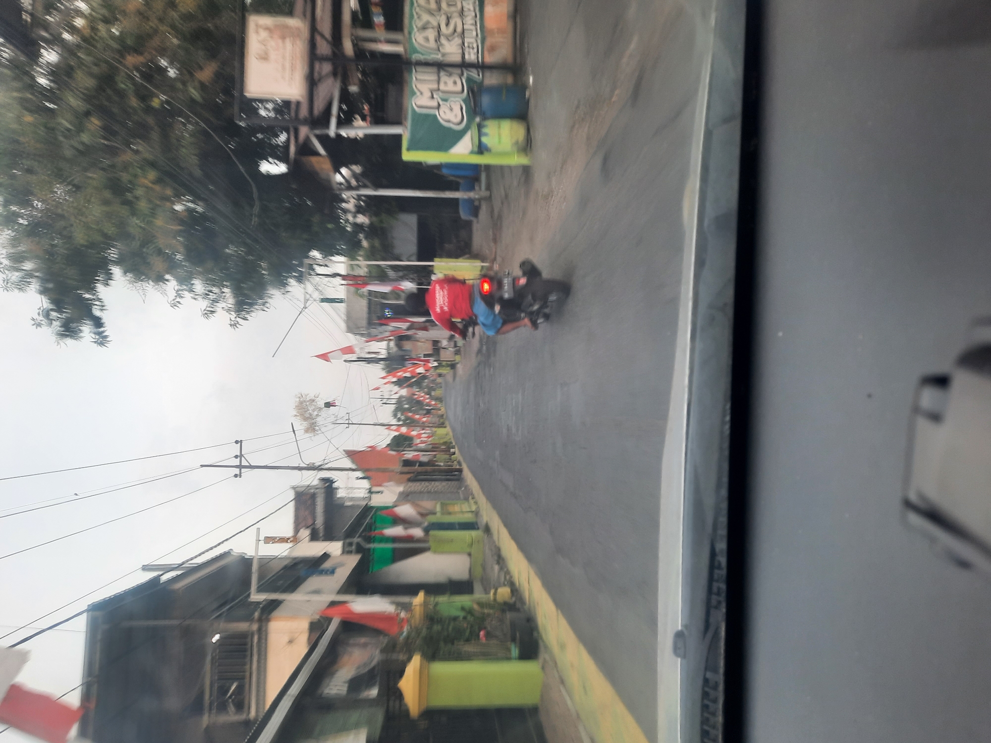 A street scene in Banjarkemantren, Indonesia, features numerous Indonesian flags lining the road, suggesting a national celebration or commemoration. The perspective is from inside a vehicle, looking out through the windshield. A man in a red shirt and blue shorts is riding a motorcycle down the street. Buildings with balconies and awnings are visible on the left side of the road, and trees and a vendor stall are on the right. The sky is overcast.