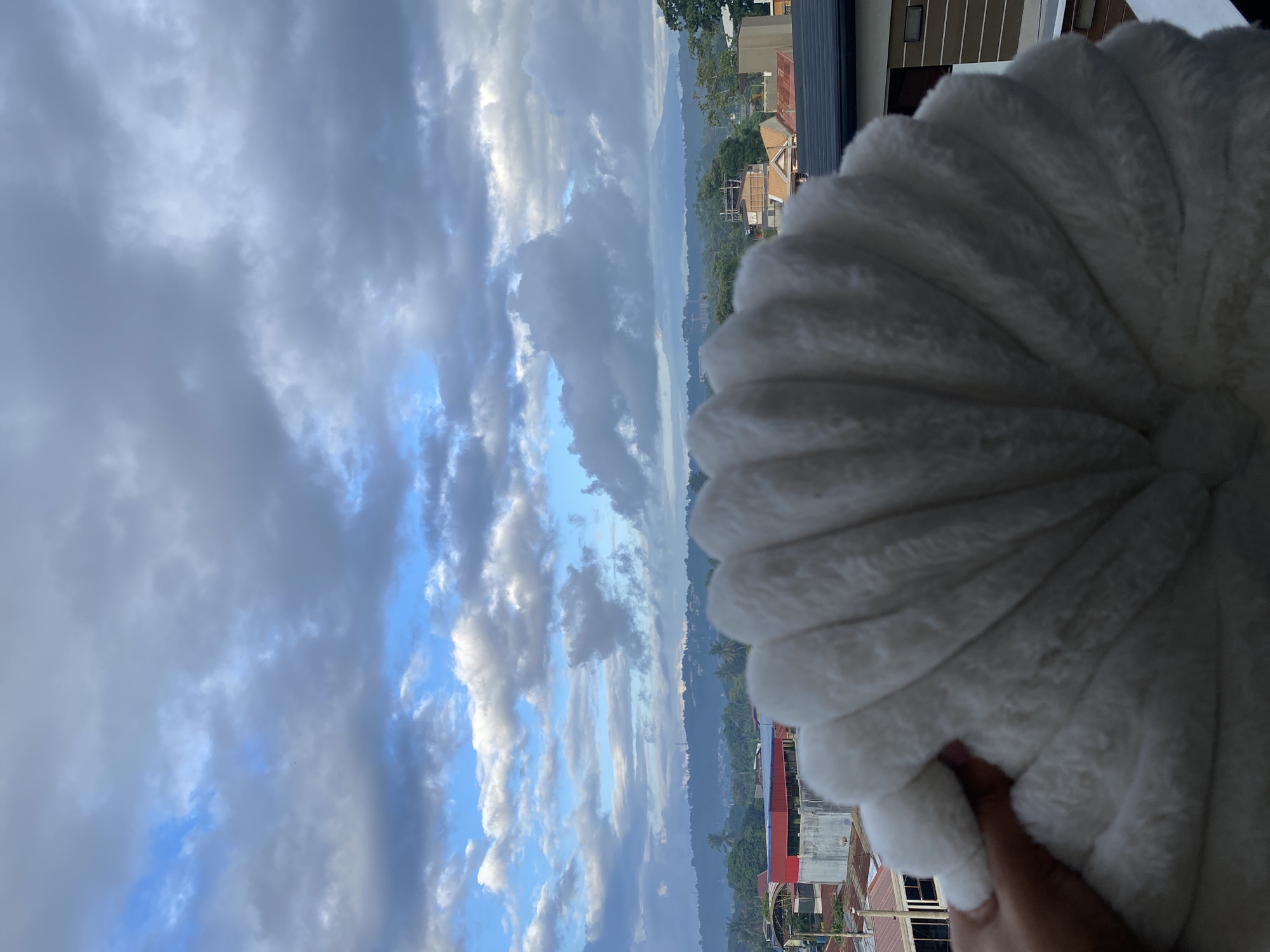 A hand holds a fluffy white object resembling a large seashell or fan in the foreground, obscuring a portion of a cityscape and a mountainous landscape under a dramatic cloudy sky. The buildings visible in the mid-ground appear to be residential or commercial structures with red and orange rooftops. Beyond the immediate urban area, rolling hills and mountains are silhouetted against the horizon. The sky is filled with a mix of dark gray and bright blue clouds, suggesting dynamic weather conditions. The scene is set in Davao City, Philippines, as indicated by the distinctive cloud formations and mountainous terrain typical of the region.