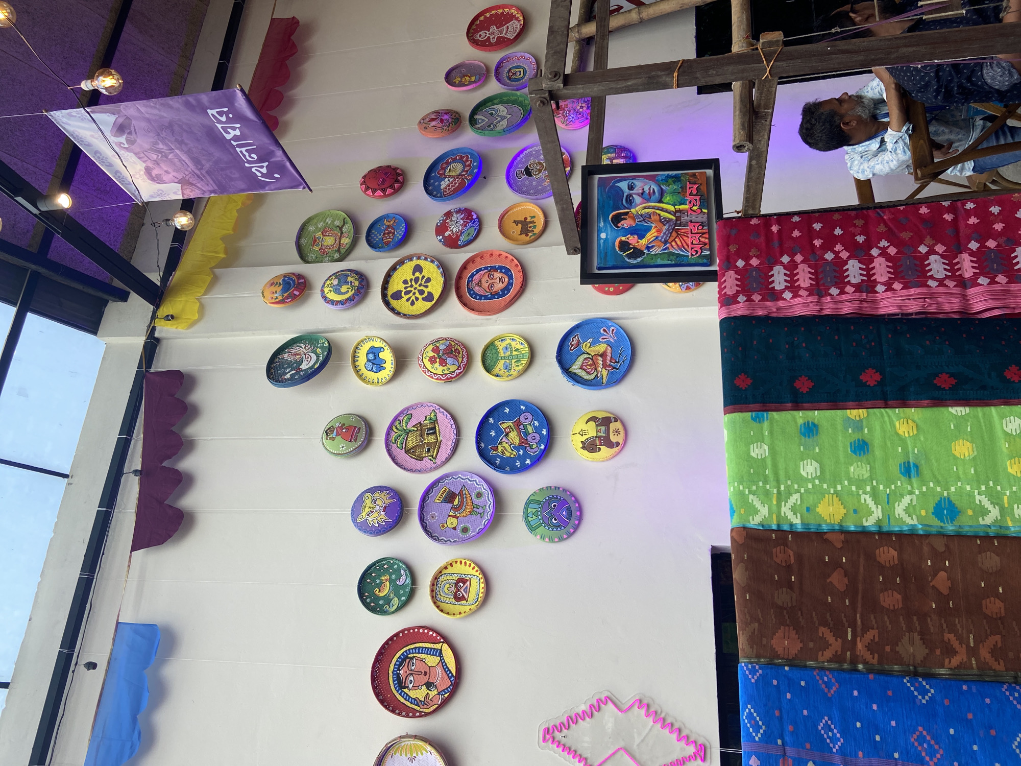 The image shows an indoor scene in Dhaka, Bangladesh, likely a shop or display area featuring traditional crafts and decorations. The wall is decorated with a variety of painted plates with folk art motifs, arranged in a decorative pattern. A framed painting depicting a scene or figures, is also visible. A wooden structure or scaffolding is present, with a person weaving on a loom, suggesting a working craft studio. Several colorful fabrics with traditional patterns hang near the loom. A purple banner or sign is visible on the left side of the wall, adorned with possibly Bengali script.