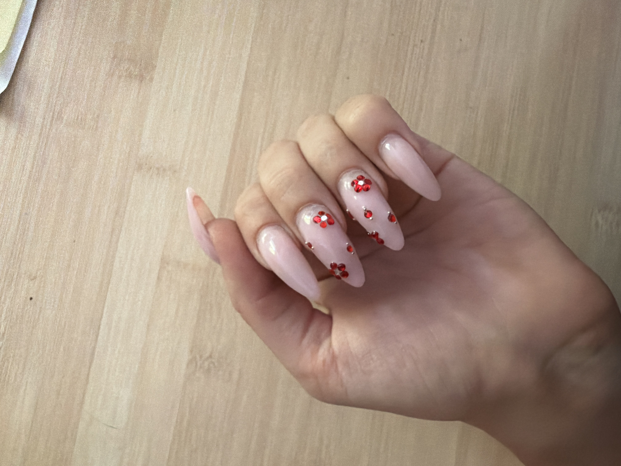 A left hand with long, light pink, almond-shaped nails is held up against a light brown wooden background. The nails are adorned with small, red, flower-shaped rhinestones and scattered silver rhinestones. The hand is relaxed, with fingers slightly curled. The lighting suggests an indoor environment. The image does not contain any information about the city of Prosena, Bulgaria.