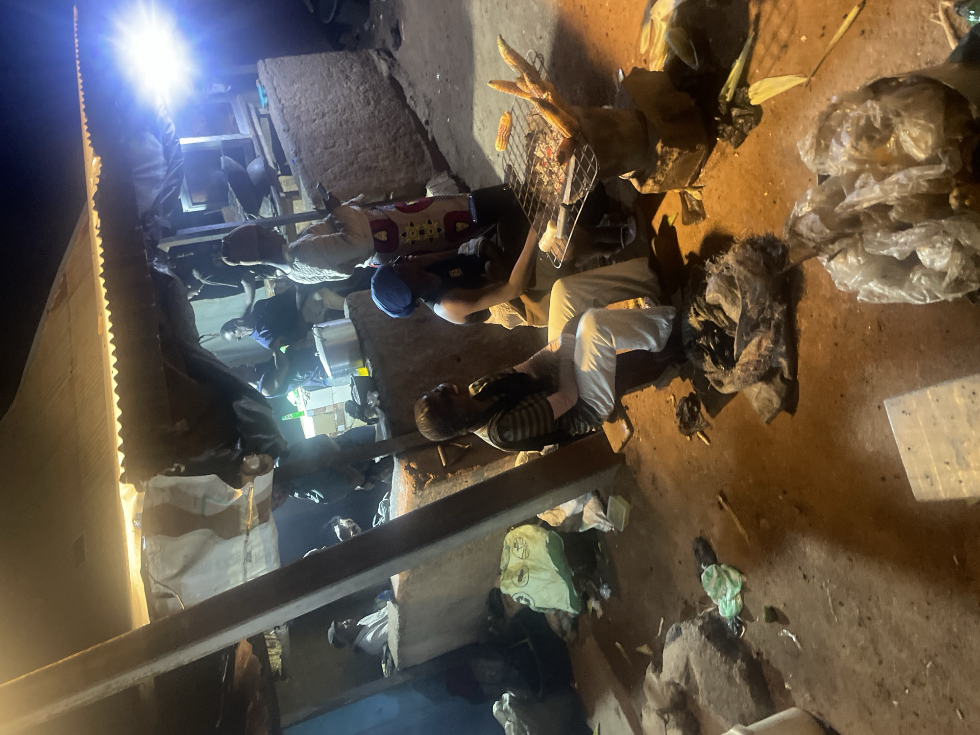 The image depicts a nighttime scene in Du, Nigeria, likely a street food stall. Several individuals are present, some engaged in food preparation and others observing. In the foreground, a person wearing a blue headscarf is tending to corn being grilled over coals. To the left, another individual sits wearing a striped shirt and white pants. In the background, other people are visible, including one by a food stall and another in a blue shirt. The environment appears to be rustic, with a corrugated metal roof overhead and a dirt ground. Various items, including cooking utensils, plastic bags, and discarded materials, are scattered around the area. A bright light illuminates the scene from the right, suggesting artificial lighting.