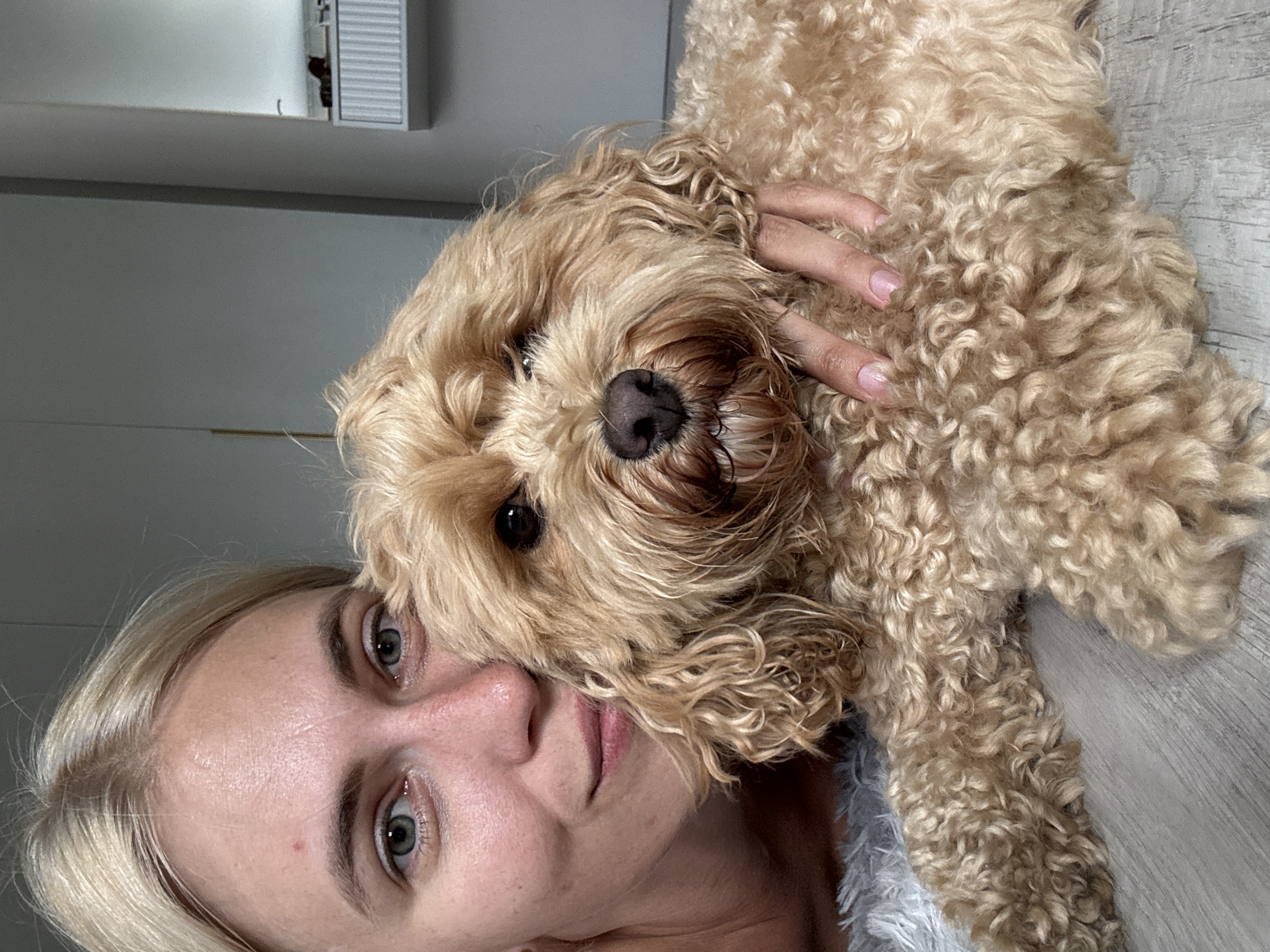 A close-up, selfie-style photograph features a fair-skinned woman with blonde hair and blue-green eyes, smiling slightly as she embraces a golden-colored, curly-haired dog. The dog's head rests against the woman's face, and her hand is visible stroking its back. They are positioned on a light-colored, textured surface, possibly a rug or blanket, with a wooden floor visible beneath. The background is out of focus, but appears to be an indoor setting with a wall and cabinetry, suggesting a domestic environment. There is no indication of the city of Saratov, Russia, in the image.