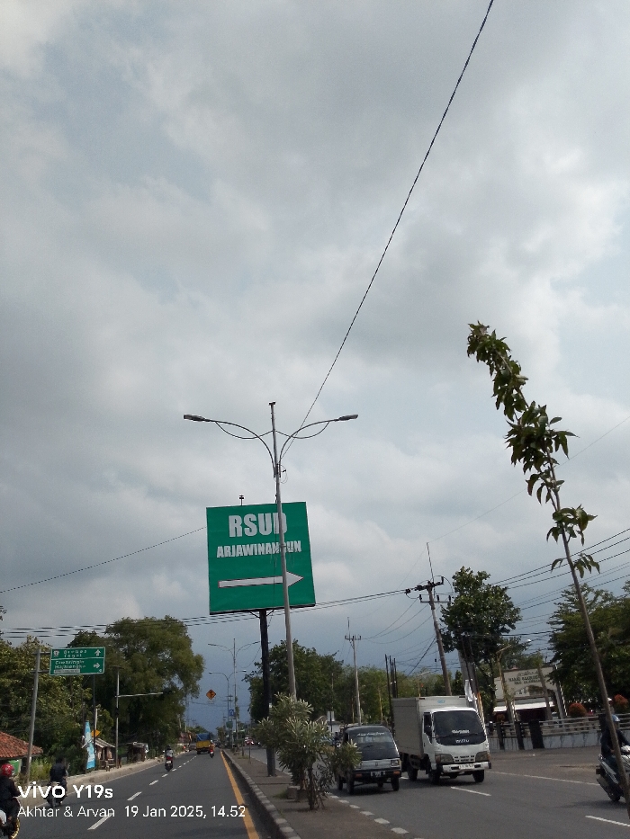 A wide-angle, eye-level photograph captures a bustling road scene in Arjawinangun, Indonesia, under an overcast sky. The road, featuring multiple lanes, stretches into the distance, bordered by trees and utility poles. The image, timestamped January 19, 2025, at 14:52, was taken with a VIVO Y19s.
In the foreground and mid-ground, various vehicles navigate the road. On the right side, a white utility truck and a smaller dark pick-up truck are visible, moving away from the viewer. Further down the road, more cars and motorcycles can be seen in both directions. A person on a motorcycle is partially visible in the bottom left corner.
A large green rectangular sign stands prominently on the right side of the road, mounted on a tall pole that also supports a modern dual-fixture street lamp. The sign reads "RSUD ARJAWINANGUN" in white text, indicating a regional general hospital, with a clear white arrow pointing to the right. On the left side of the road, a smaller green sign with white text lists several destinations, including "Cirebon," "Jakarta," "Brebes," "Kuningan," and "Majalengka," along with directional arrows.
The sky is heavily clouded with large grey and white clouds, creating diffuse lighting characteristic of an overcast day. Numerous utility poles with intertwined electrical wires span across the frame, with one prominent wire running diagonally across the upper part of the image. Lush green trees and smaller bushes line both sides of the road and the central median. A distinctive, slender tree branch with green leaves extends into the upper right corner of the frame, adding a natural element. The scene depicts a typical, busy urban or semi-urban roadway.