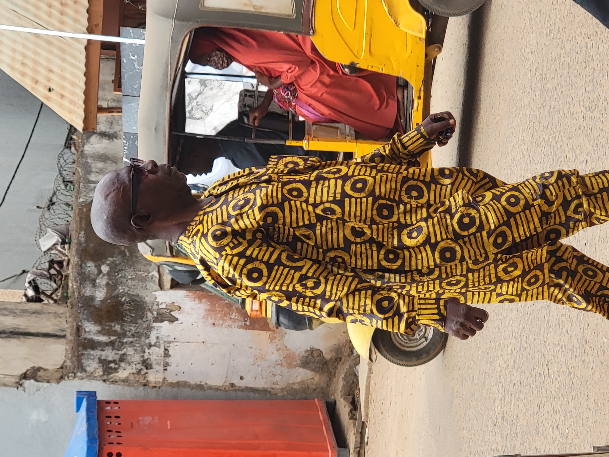 A middle-aged man with dark skin and a bald head, wearing glasses and a yellow and black patterned shirt and pants, is walking next to a yellow auto rickshaw (tuk-tuk) on a paved street. The tuk-tuk contains two passengers; one is wearing red attire. The background includes a building with a corrugated metal roof and weathered walls. A red and blue storage container sits near the building. The location is Maiburiji, Nigeria.