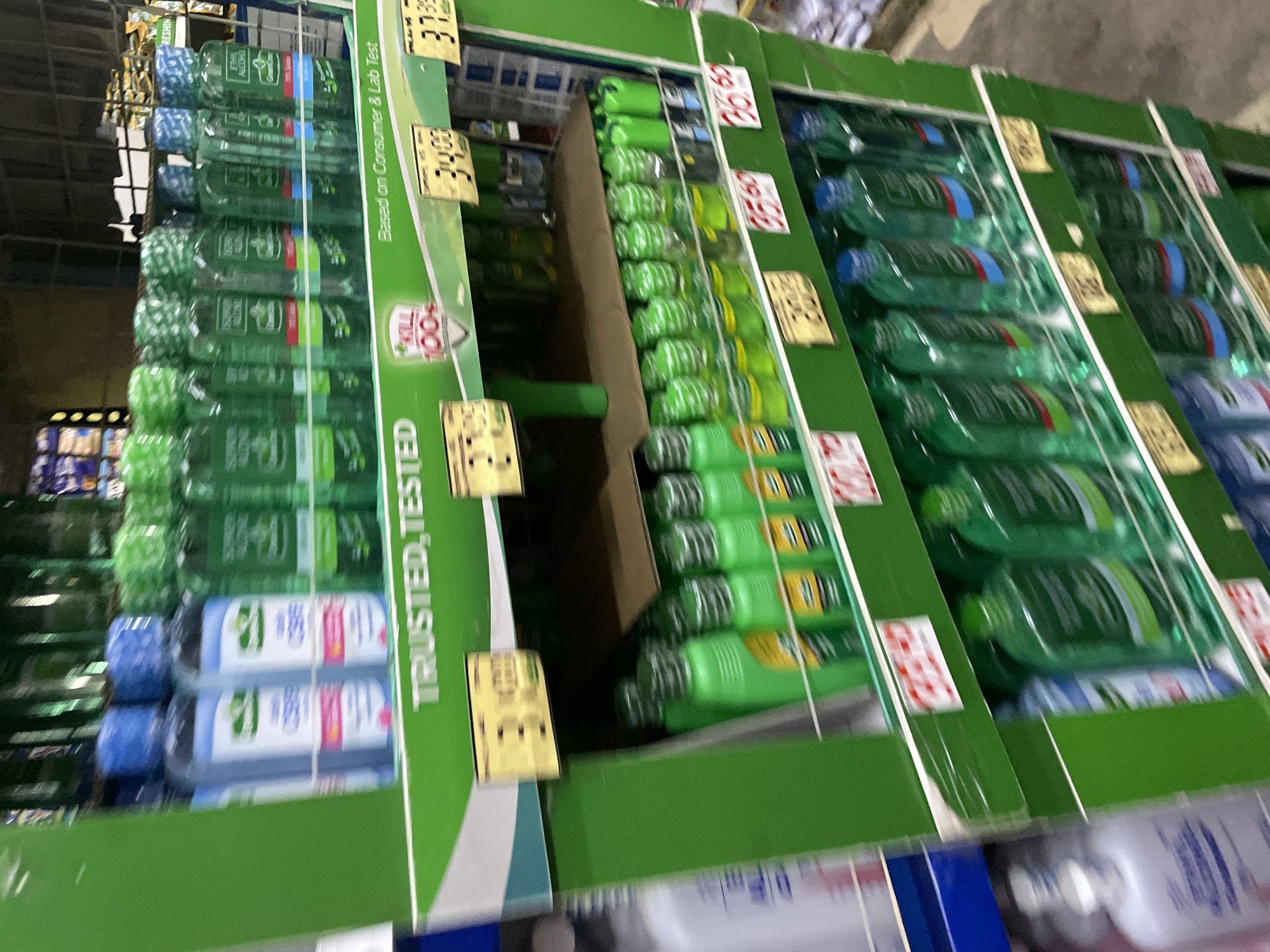 The image depicts a multi-tiered retail display stand, predominantly green and constructed of cardboard, located within a supermarket or pharmacy setting in Davao City, Philippines. The stand is filled with numerous plastic bottles of hand sanitizer or rubbing alcohol.
The top shelf features two distinct types of bottled products. On the left, several light blue translucent bottles with white labels and blue caps are arranged. To their right, a row of green translucent bottles with green caps and labels, some clearly visible as "ETHYL ALCOHOL" with a "Green Cross" logo, are displayed. The shelf edge bears the text "TRUSTED, TESTED" and a shield-shaped logo with "KILL 100+". Yellow price tags are affixed to the shelf, showing amounts such as "34.00" and "37.99". The phrase "Based on Consumer & Lab Test" is also visible on the shelf.
The middle shelf, partially obscured, holds green bottles, many with yellow labels, arranged in rows behind a cardboard divider. Another yellow price tag displays "35.60" and "36.50" on this level.
The bottom visible shelf displays more green translucent bottles, characterized by blue or red banded labels and blue caps. Additional yellow price tags are visible, with prices such as "33.99" and "32.99" (partially obscured).
To the far left and right edges of the frame, glimpses of other retail shelves or products, including white and blue bottles, are visible but out of focus. The background shows blurry shelves stocked with various other items. The floor is visible at the bottom right, appearing as a dark, possibly concrete, surface. No individuals are present in the frame.