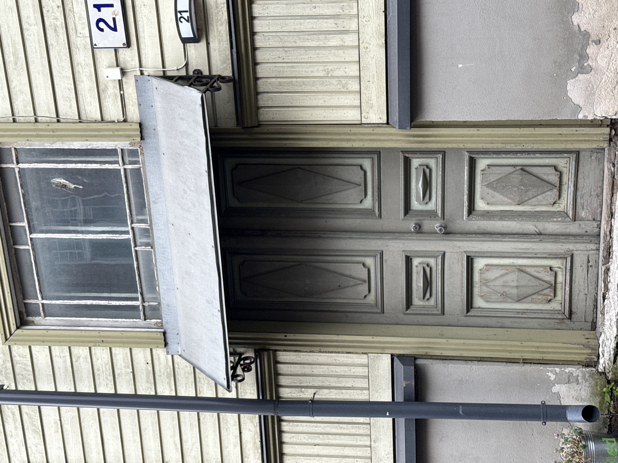 The image shows the exterior of a building with a green double door and a window above it. A dark gray downspout runs vertically along the left side of the image. Above the door, there is a metal awning. The building's facade is clad in horizontal wooden planks painted a light yellow-green. To the right of the door, on the wooden facade, are two house number signs, both displaying "21". One sign is larger and placed higher, while the other is smaller and located below it. A small white box is mounted on the wall between the window and the upper house number. The door itself is old and weathered, with decorative diamond-shaped panels. The window above the door has multiple panes with white mullions. In the lower left corner, a metal bucket with some small plants is visible, and a drainpipe extends from the downspout towards the ground. The scene appears to be a street view in Tallinn, Estonia.