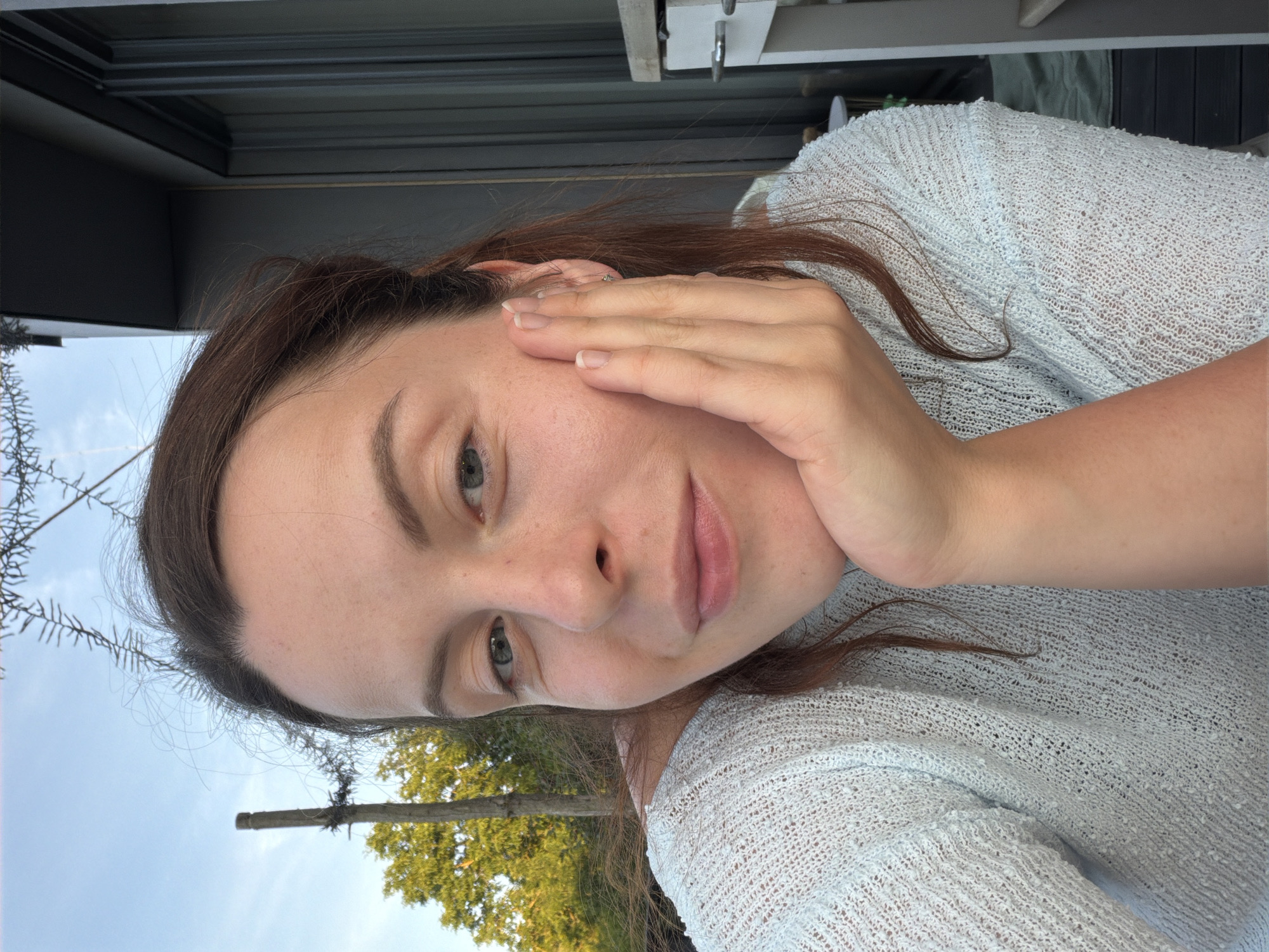 A woman is captured in a close-up selfie outdoors, with her face resting on her hand. She has brown hair and is wearing a light blue, textured sweater. The background shows a blue sky with some tree branches and a building. The image appears to be taken during the daytime.