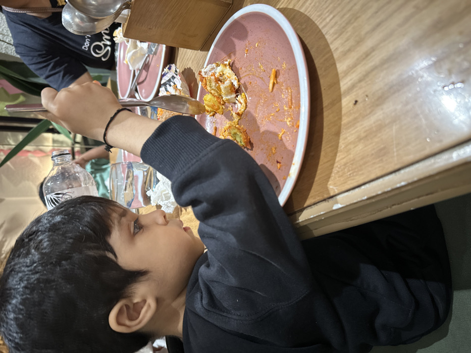 An indoor scene in a restaurant in Kalyan-Dombivli, India, depicts a young child seated at a light-colored wooden table. The child, with dark hair, is seen in profile from the chest up, facing right, wearing a black long-sleeved garment. A black thread bracelet is visible on his right wrist. He holds a silver-colored spoon in his right hand, positioned over a round pink plate with a white rim, which contains remnants of a multi-colored food item (white, green, orange/red) and reddish spice residue. To the left of the plate, a clear glass of water with condensation and a crumpled white paper napkin are on the table. In the background, a person with a light green shirt is visible holding a clear plastic water bottle to their mouth. To the right, another person's arm and upper torso are visible, wearing a dark t-shirt with partially legible white text, and orange-painted fingernails. A wooden caddy holding additional silver utensils is situated further right on the table. Other similar pink plates, mostly cleared, are also visible. The background includes blurred green foliage, suggesting decorative plants, and soft ambient lighting.
