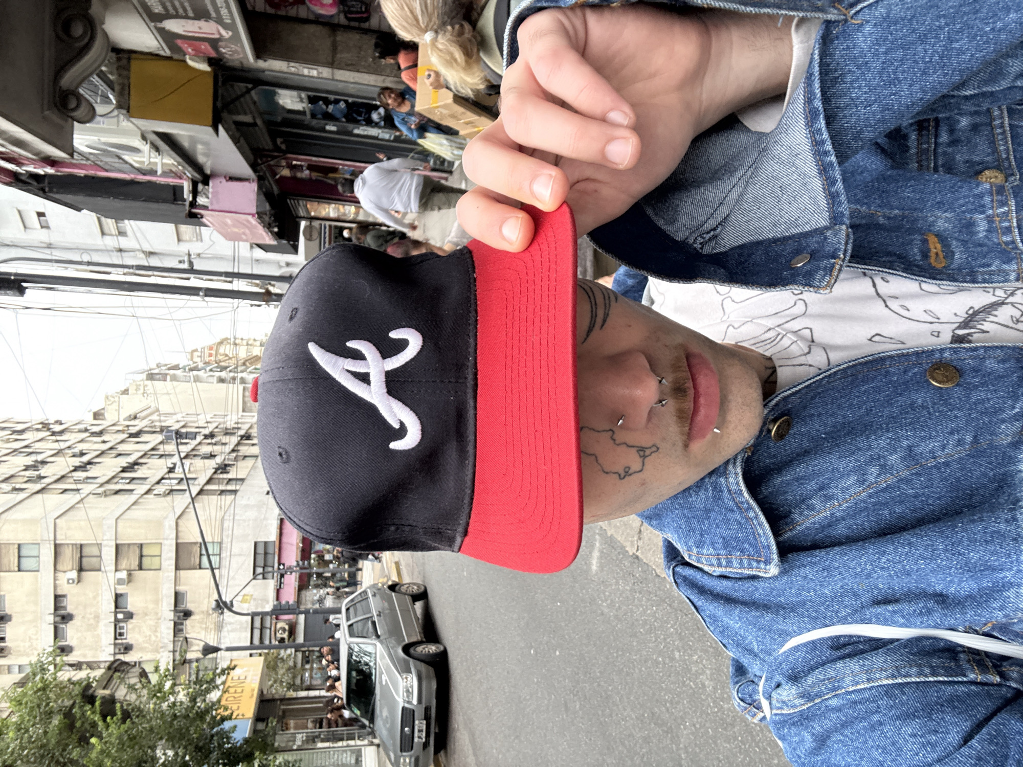 A person wearing a denim jacket and a black baseball cap with a white logo and red brim is holding the brim of the cap. The person has piercings on their face and a tattoo on their cheek. The background shows a street scene in Buenos Aires, Argentina. Buildings, including one with multiple windows and air conditioning units, are visible, along with streetlights, trees, and a parked gray car. Pedestrians are seen on the sidewalk. A sign reading "IRENE" is partially visible.