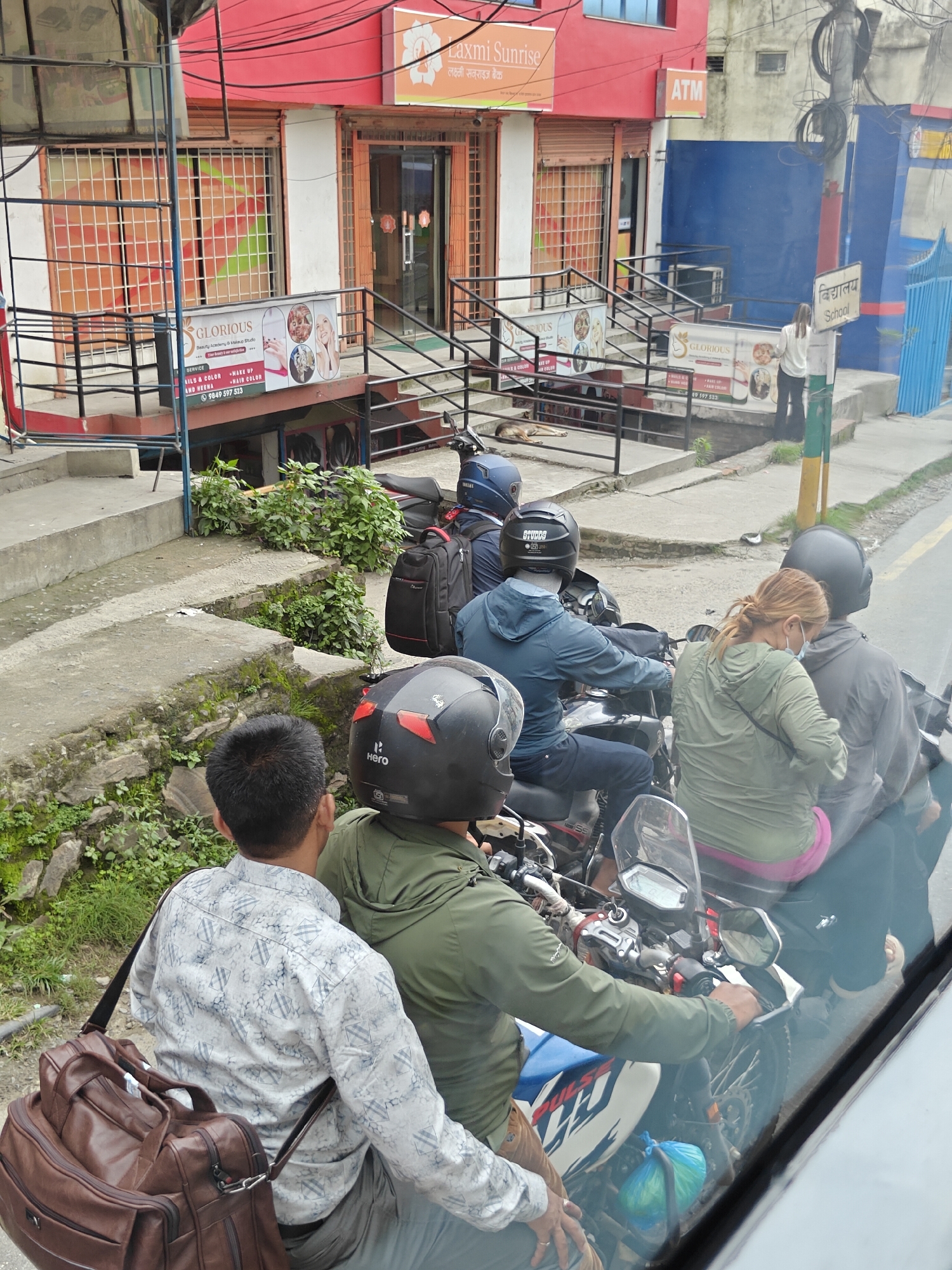 In Chandragiri, Nepal, a traffic scene unfolds on a paved road, viewed from inside a vehicle. A group of motorcyclists are stopped at what appears to be a junction or traffic light. The central focus is a motorcycle carrying three people. The driver, a man in a green jacket and a black helmet, operates the vehicle. Behind him, a woman in a green jacket and pink pants sits with a light grey jacket draped over her shoulders, her hair pulled back in a ponytail. The third passenger, a man in a grey jacket and helmet, is partially obscured.
To the left of this group, another motorcycle is visible with a driver wearing a blue jacket and helmet. A brown leather bag is slung over the shoulder of a man in a patterned white shirt, who is seated on the first motorcycle, suggesting he is also part of the group.
The background features commercial establishments. A red building with various signage is prominent, including "Laxmi Sunrise" and "Glorious Beauty Academy & Makeup Studio". An ATM machine is also visible. Further down the street, a blue structure, possibly a school or community center, is present with a sign that reads "School" in Nepali. A young dog is seen resting on the concrete steps leading up to one of the buildings. The overall environment suggests a busy urban area with a mix of commercial and public service spaces. The weather appears overcast, contributing to the muted lighting of the scene.