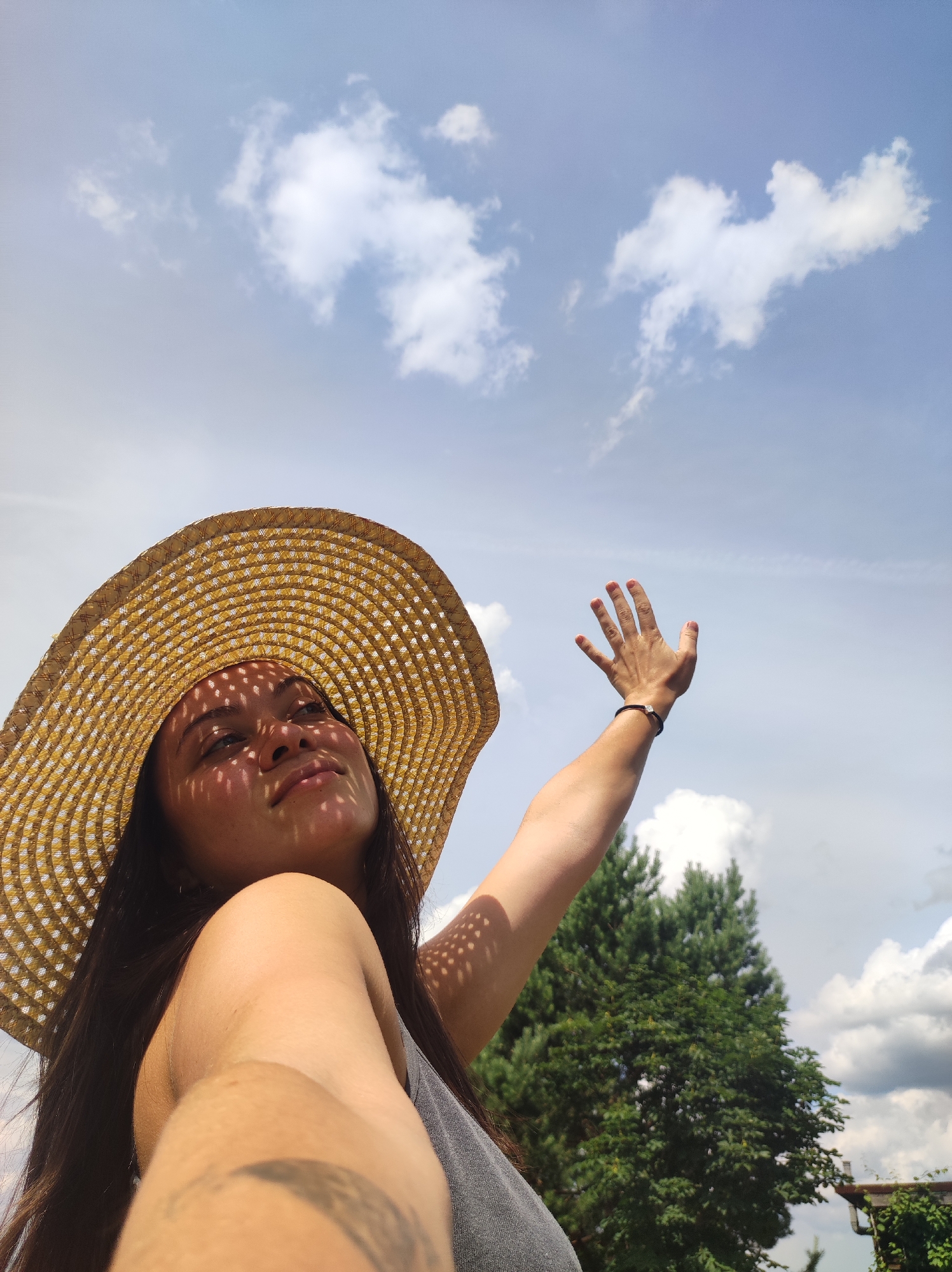 The media file features a young woman in a close-up, low-angle shot, with the sky and trees as the backdrop. She is wearing a wide-brimmed straw hat that casts a patterned shadow on her face, obscuring parts of her features while highlighting her eyes and nose. Her right arm is raised, hand open, reaching towards the sky. The woman has long, dark hair and is wearing a grey top. A faint tattoo is visible on her arm.
The scene appears to be outdoors, with lush green trees visible in the background. The sky is a bright blue with scattered white clouds, suggesting a sunny day, likely mid-morning or afternoon given the lighting. The overall impression is one of a relaxed, summery atmosphere. The woman's pose and gaze upwards suggest a moment of enjoyment or contemplation in the sunlight. There is no visible text in the media file. The location context is Фоки, Russia.